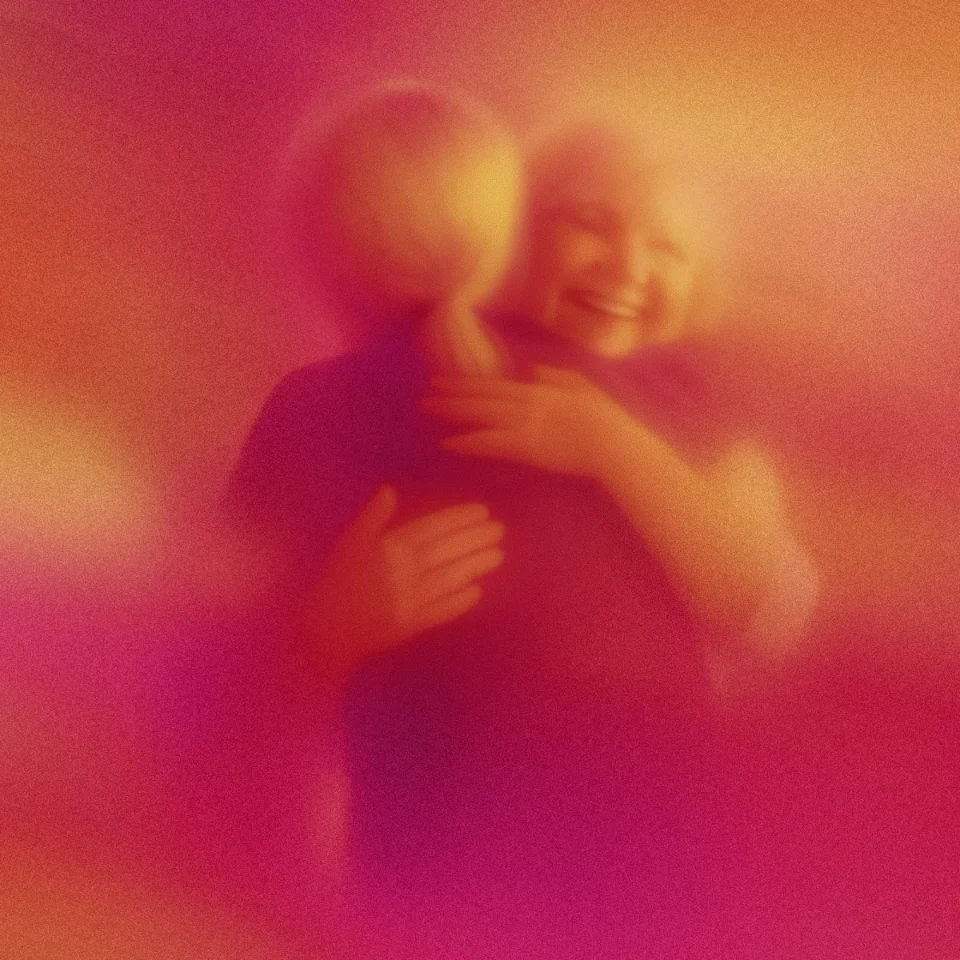 Two people embracing warmly, shown with a soft, blurred effect in warm pink and orange tones.