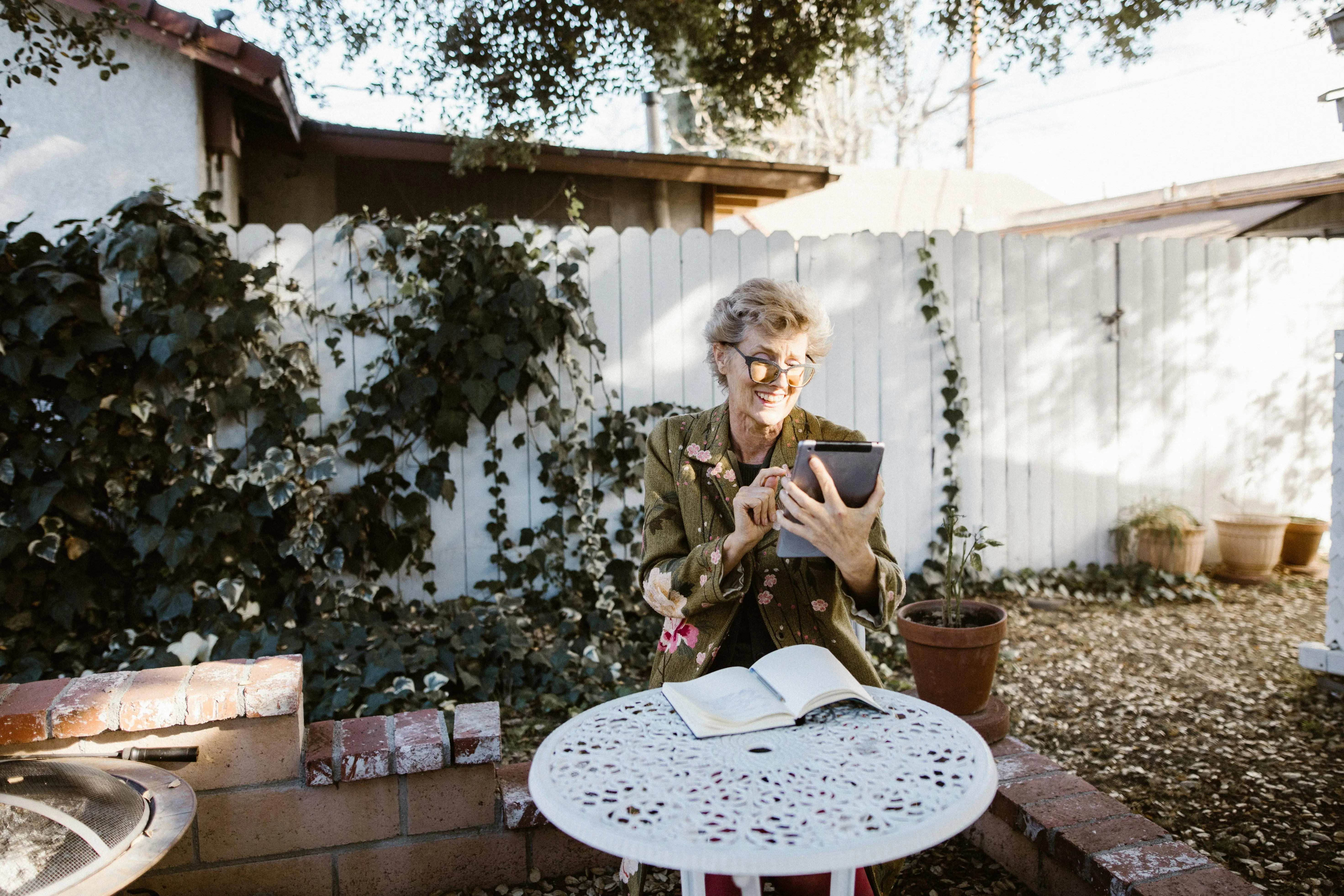 Elderly woman sitting outdoors at a white wrought iron table, smiling while using a tablet with an open notebook in front of her.