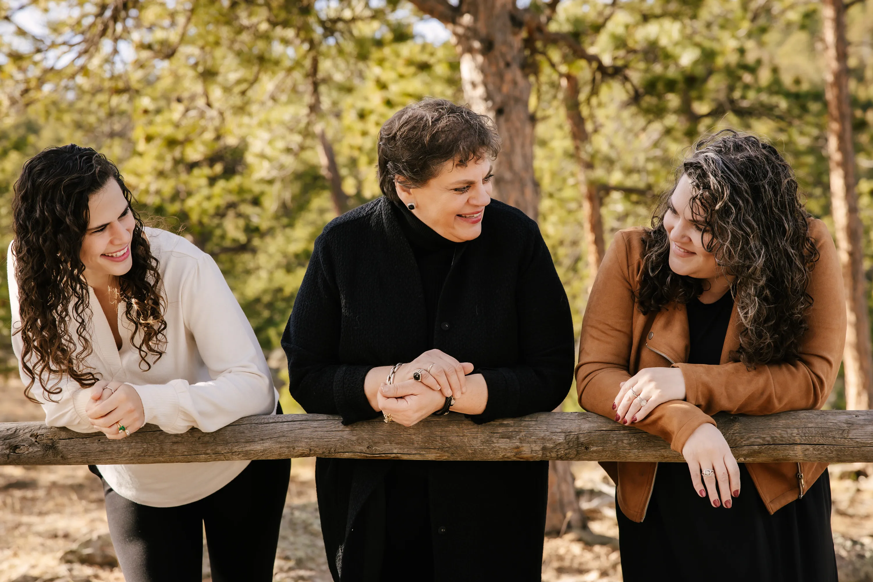 Three women leaning on a wooden fence outdoors, smiling and looking at each other amidst trees.