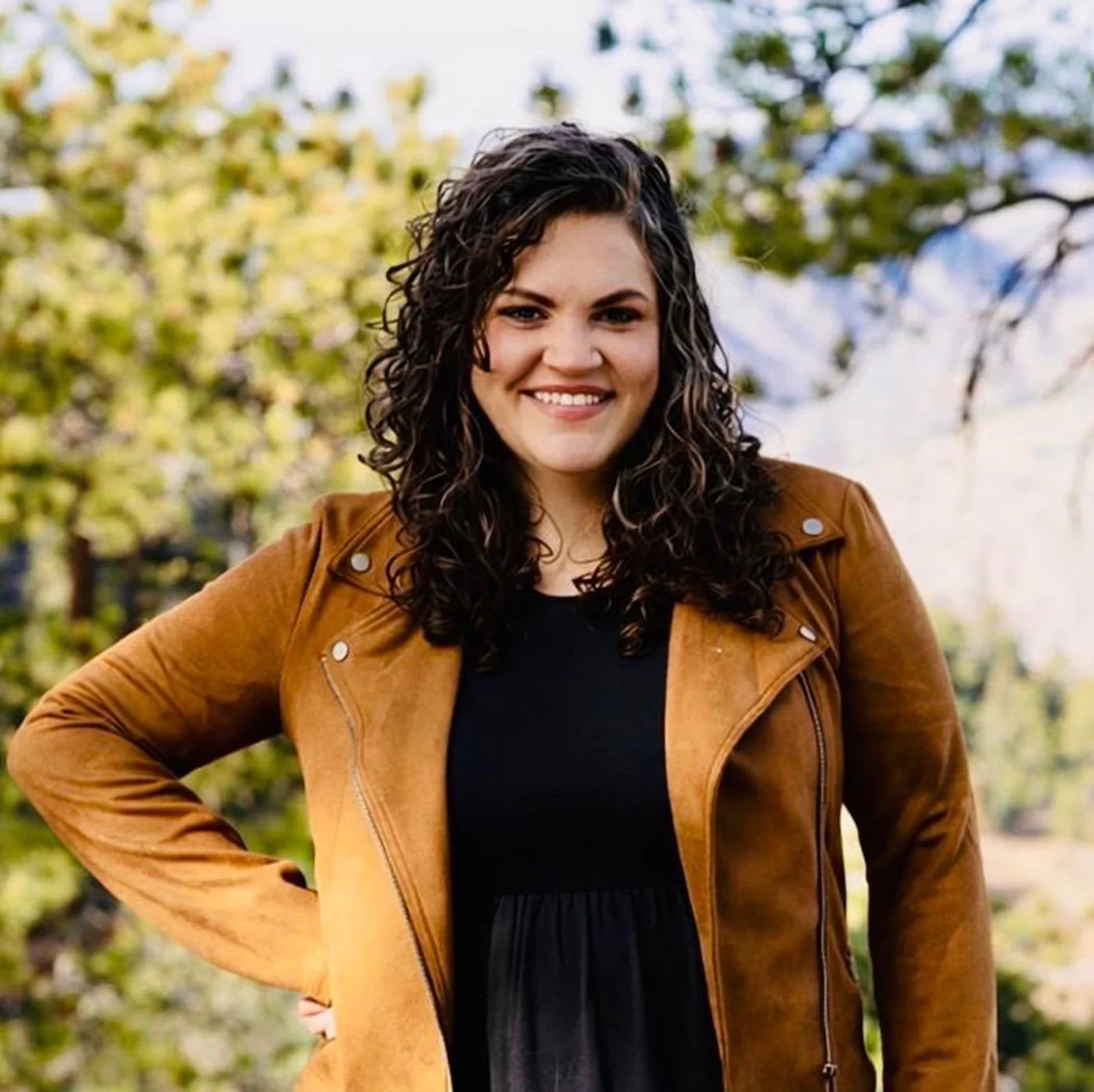 Smiling woman with curly hair wearing a brown jacket and black top outdoors with trees in the background.