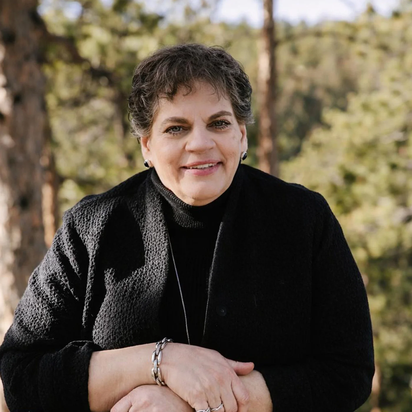 Smiling woman with short curly hair wearing a black textured sweater and silver bracelet, posing outdoors with blurred greenery background.
