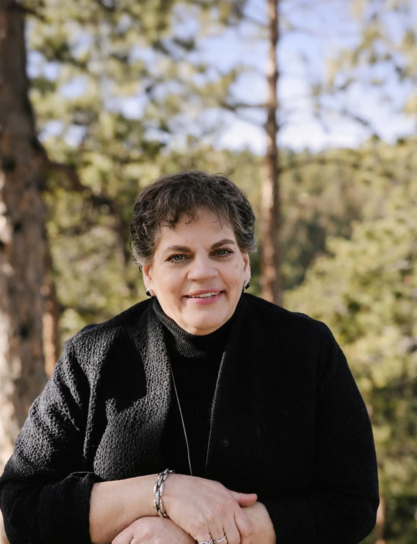 Smiling woman with short curly hair wearing a black sweater outdoors with trees in the background.
