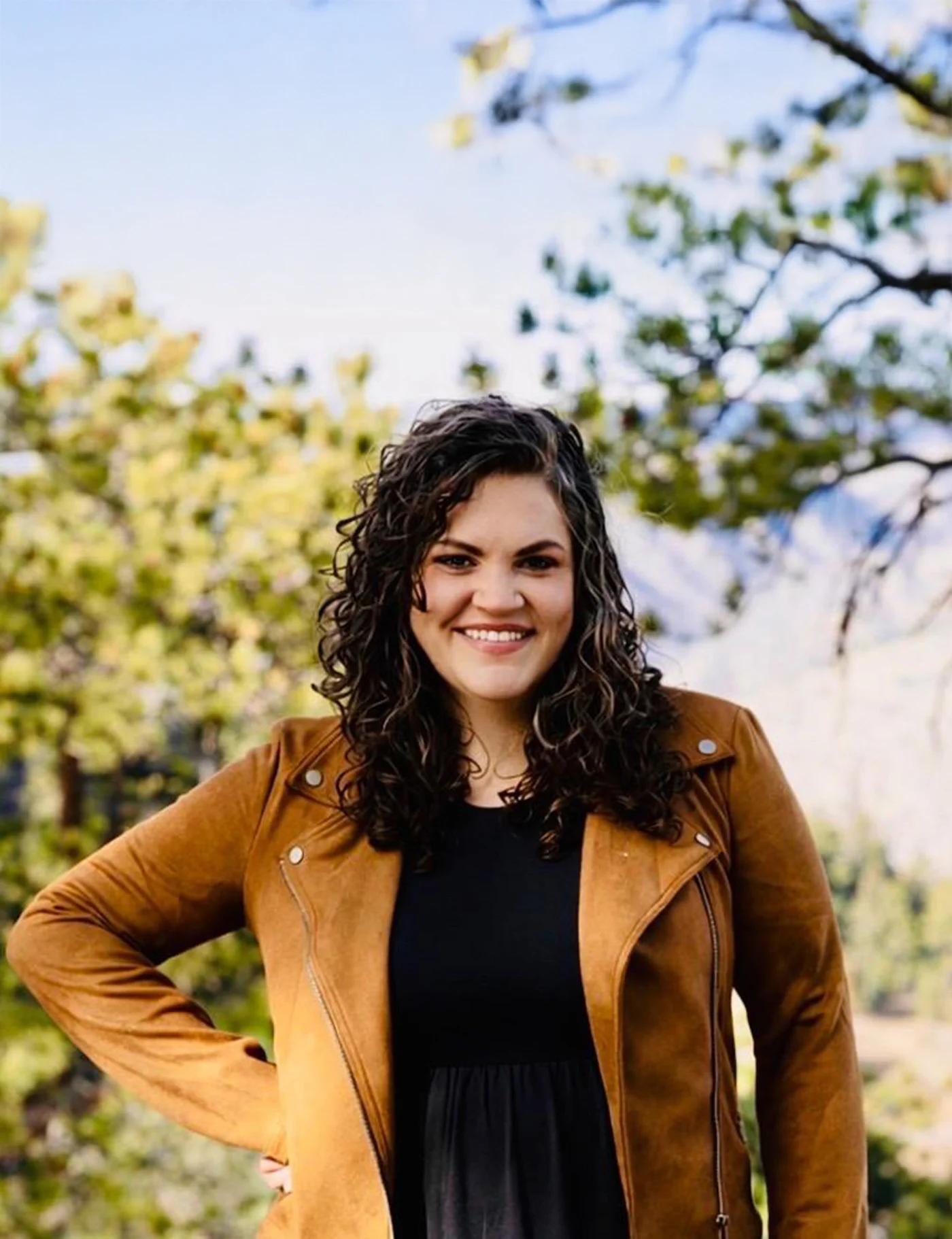 Smiling woman with curly dark hair wearing a tan jacket and black dress outdoors with trees in the background.