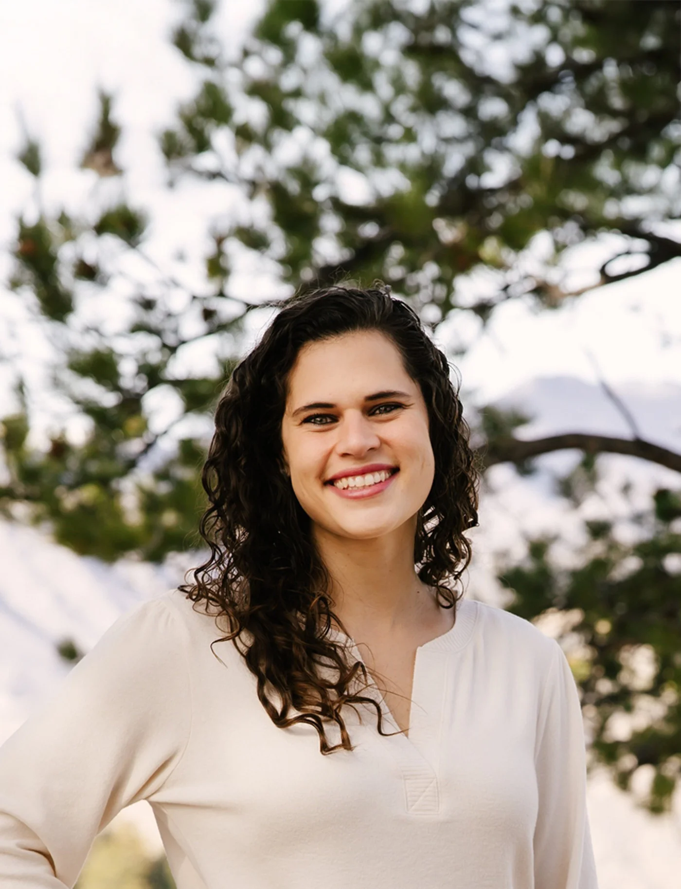 Smiling woman with curly dark hair wearing a white top standing outdoors with trees in the background.
