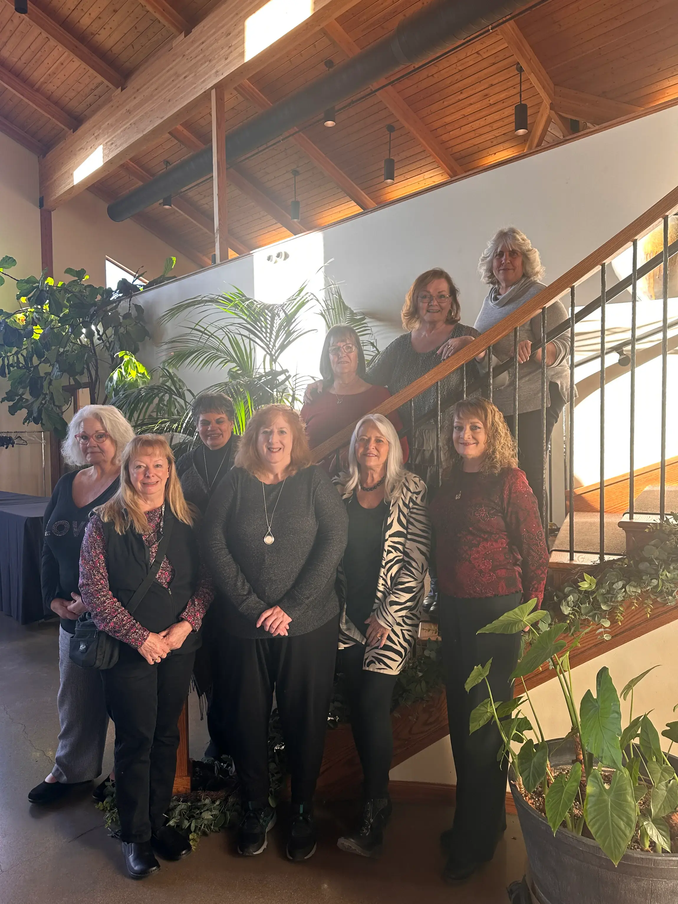 Group of nine women posing together indoors by a staircase with plants and wooden ceiling beams.