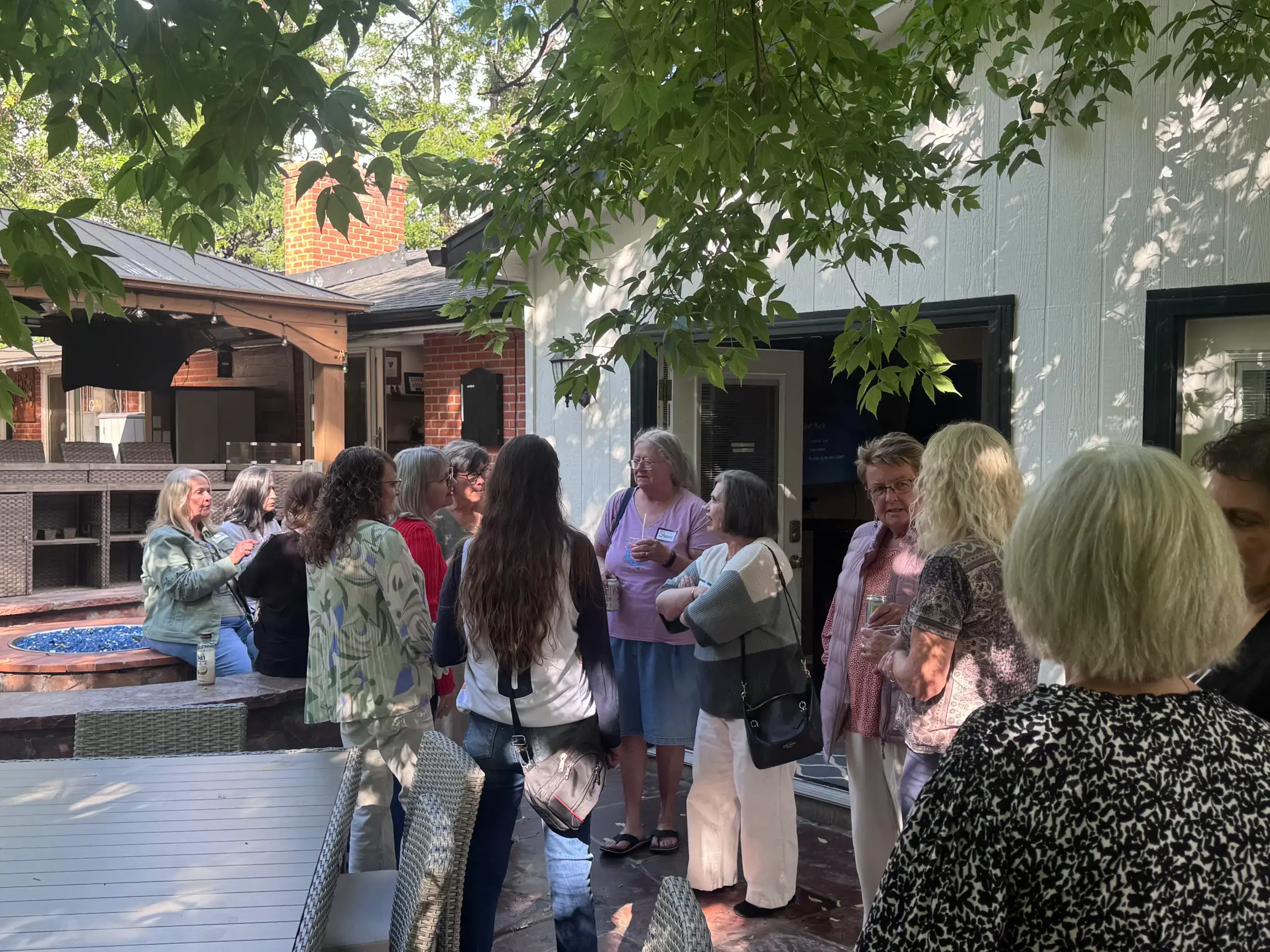 Group of women socializing outdoors on a patio shaded by tree branches near a white building.