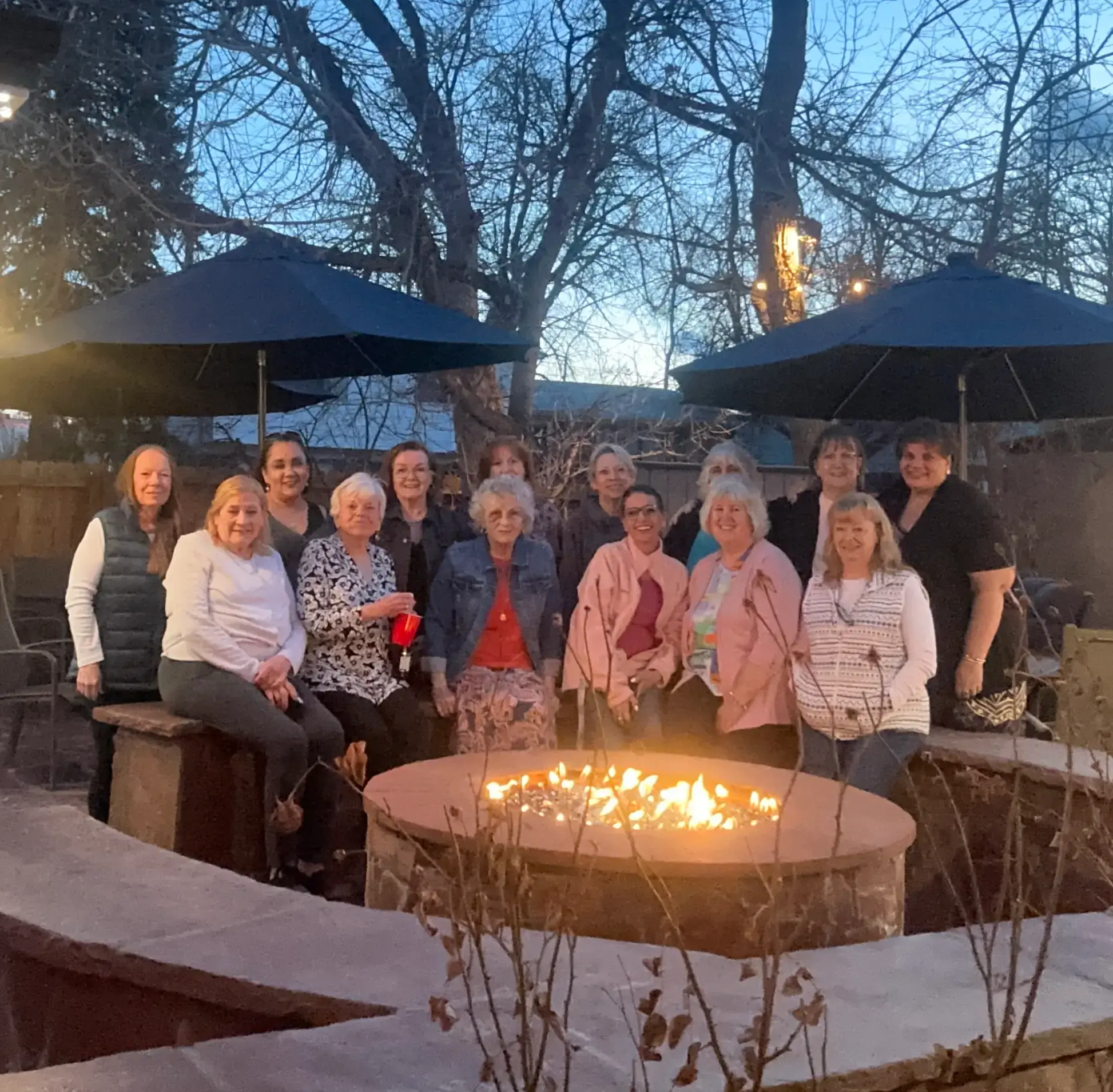 Group of fourteen women gathered around a lit outdoor fire pit at dusk with bare trees and patio umbrellas in the background.