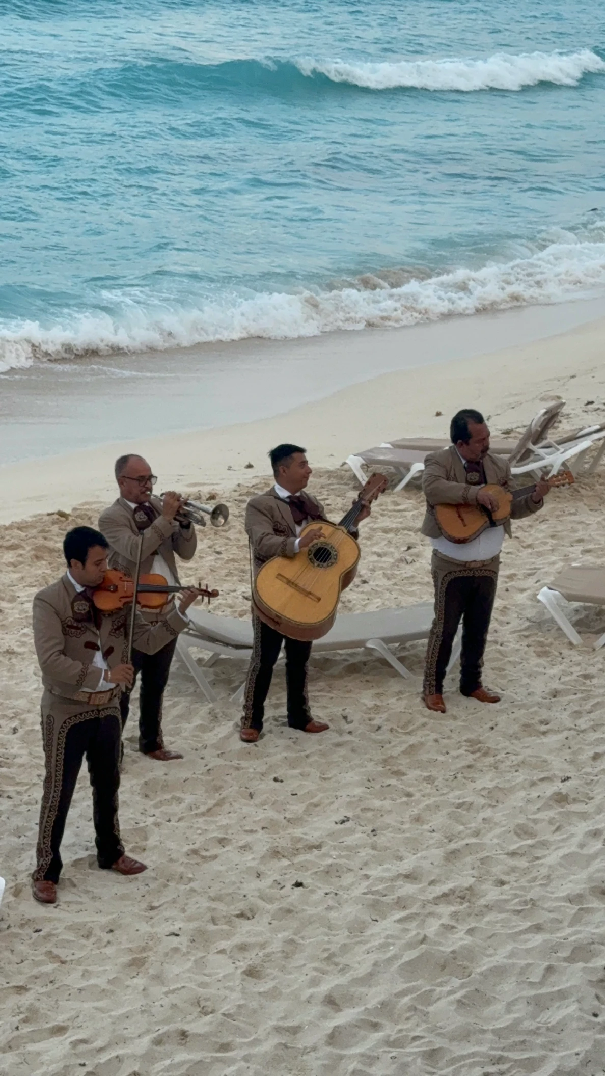 Four mariachi musicians playing instruments on a sandy beach near ocean waves.