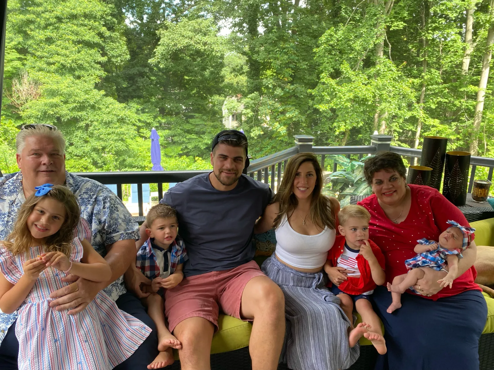 A multi-generational family sitting together on a porch sofa, surrounded by lush green trees.