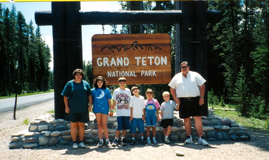 Group of six people, including four children and two adults, standing in front of a large wooden Grand Teton National Park sign with trees in the background.