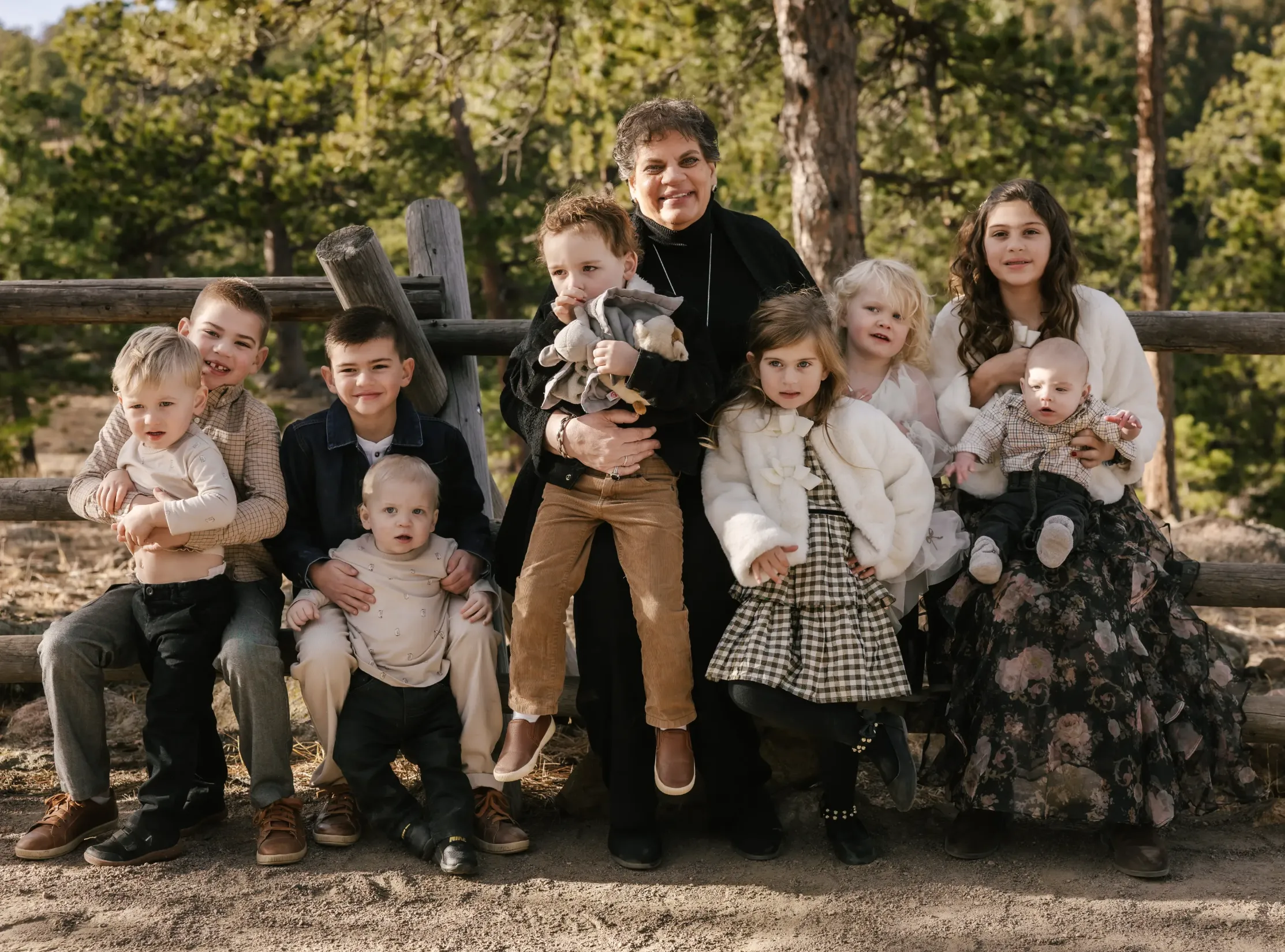 Elderly woman sitting on a wooden fence outdoors surrounded by nine children of various ages, all dressed in autumn clothing.