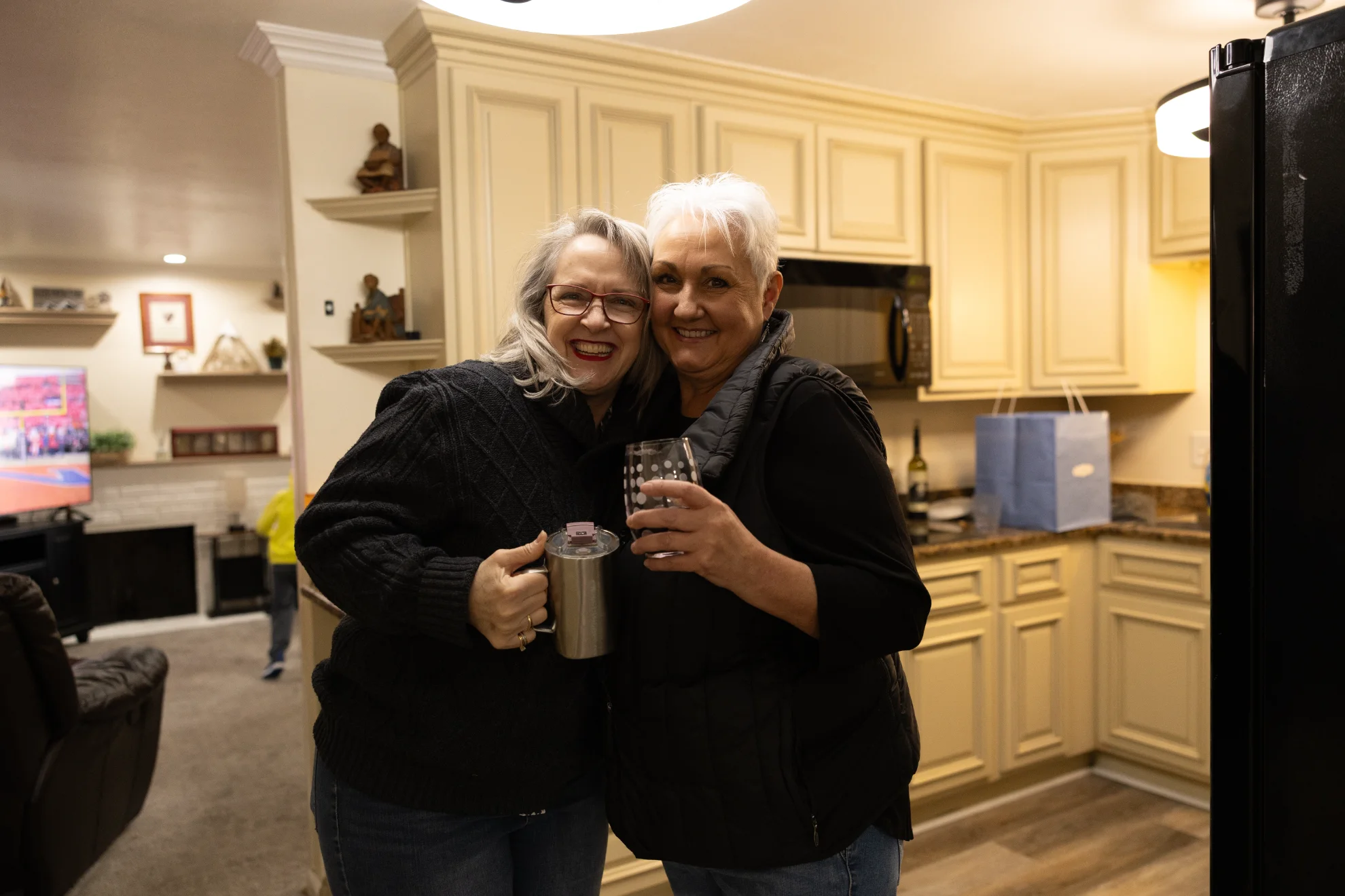 Two smiling older women hugging and holding drinks in a warmly lit kitchen.