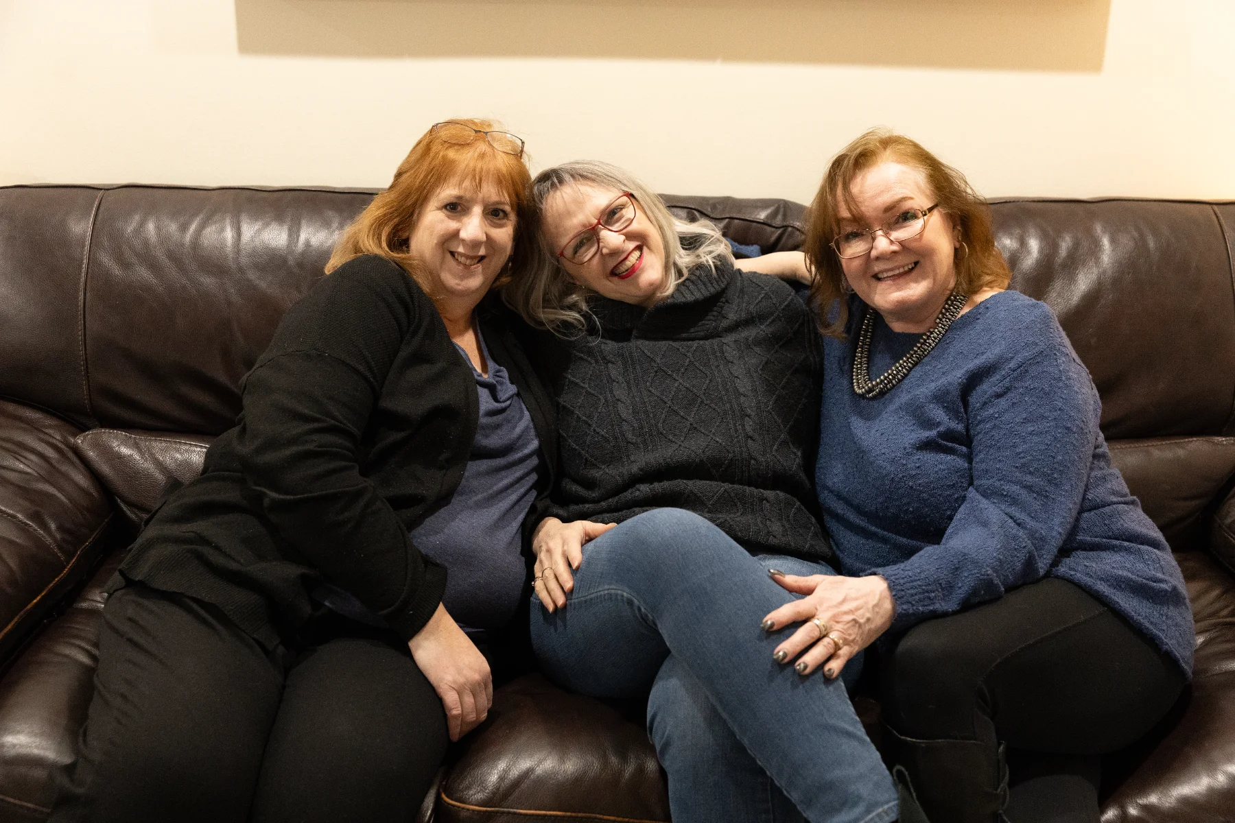 Three smiling middle-aged women sitting closely together on a dark leather couch.