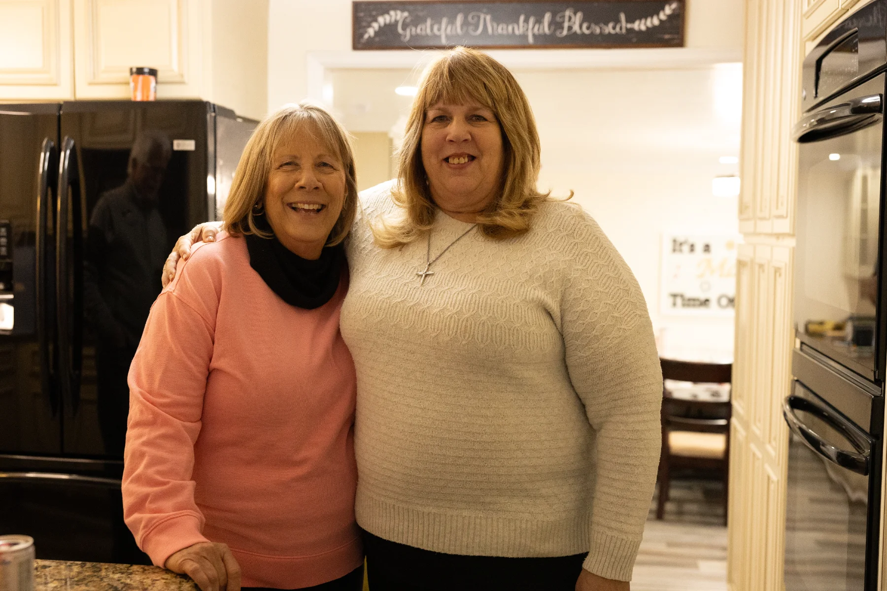 Two women smiling and embracing each other in a warmly lit kitchen with a sign reading 'Grateful Thankful Blessed' in the background.