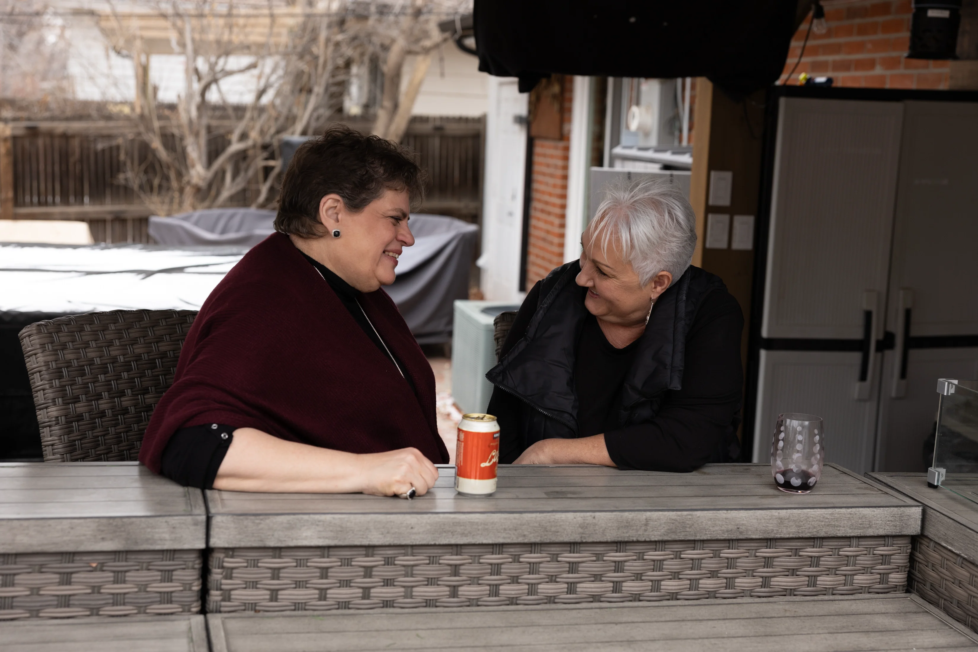 Two older women smiling and talking while sitting outdoors at a wicker patio table with drinks.