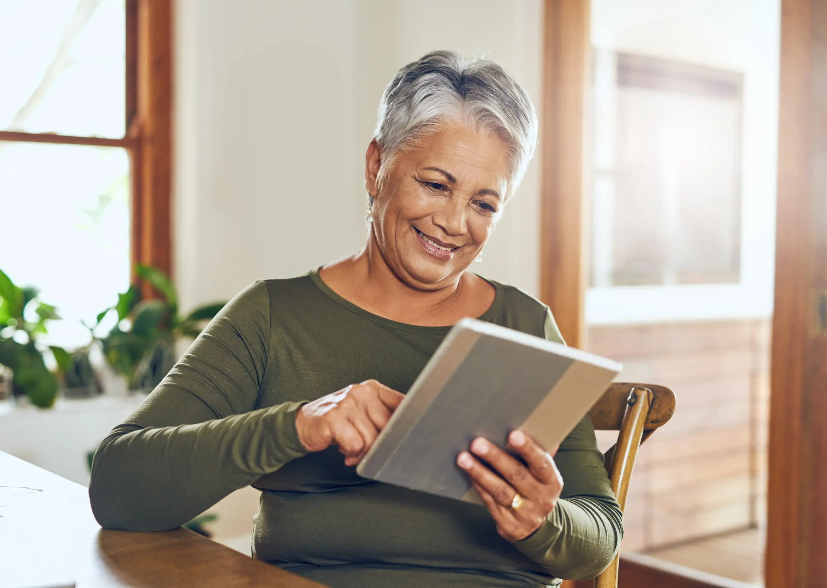 Smiling elderly woman using a digital tablet while sitting at a wooden table in a bright room.