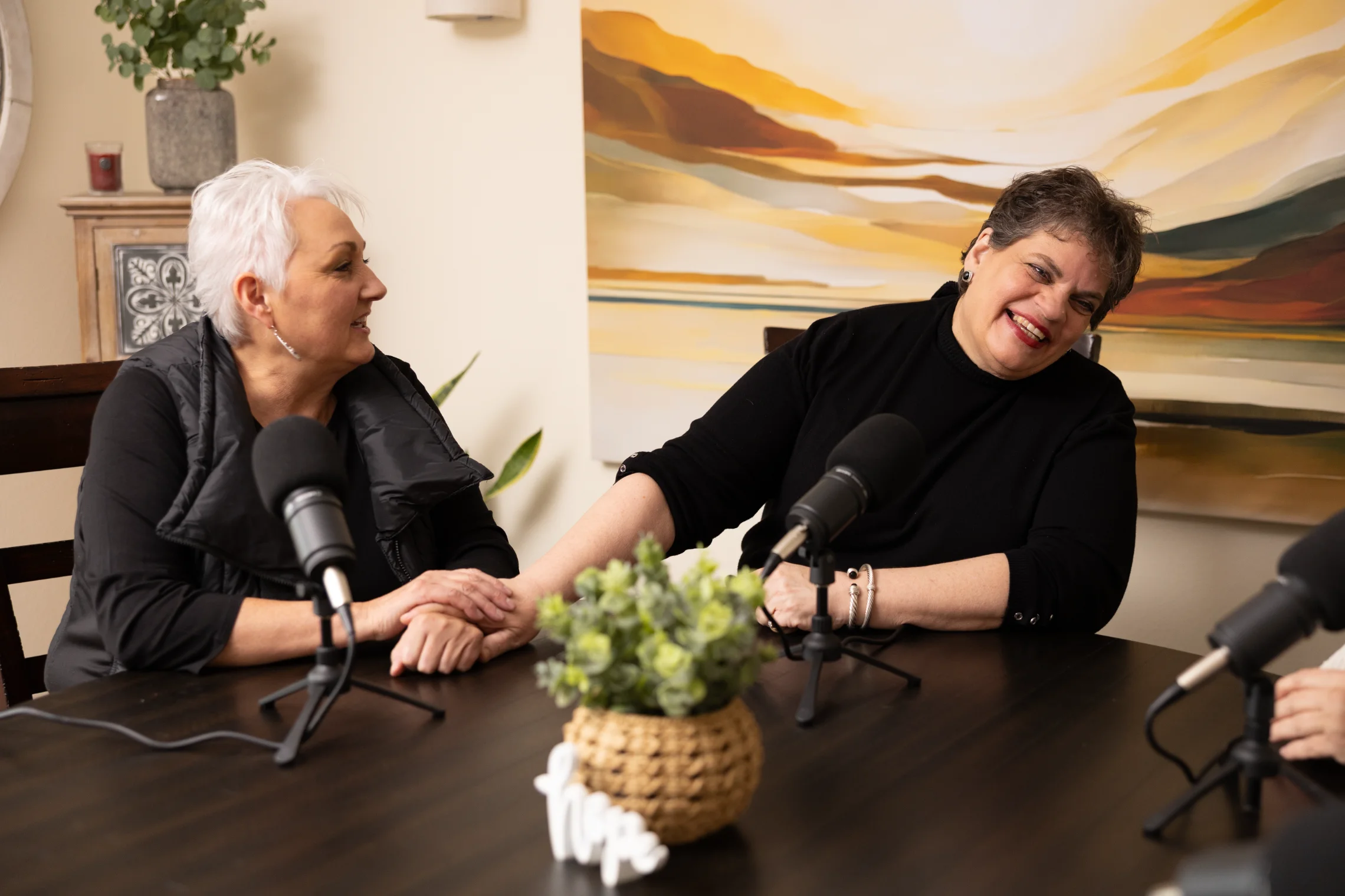 Two women sitting at a table with microphones, holding hands and smiling during a podcast or interview.