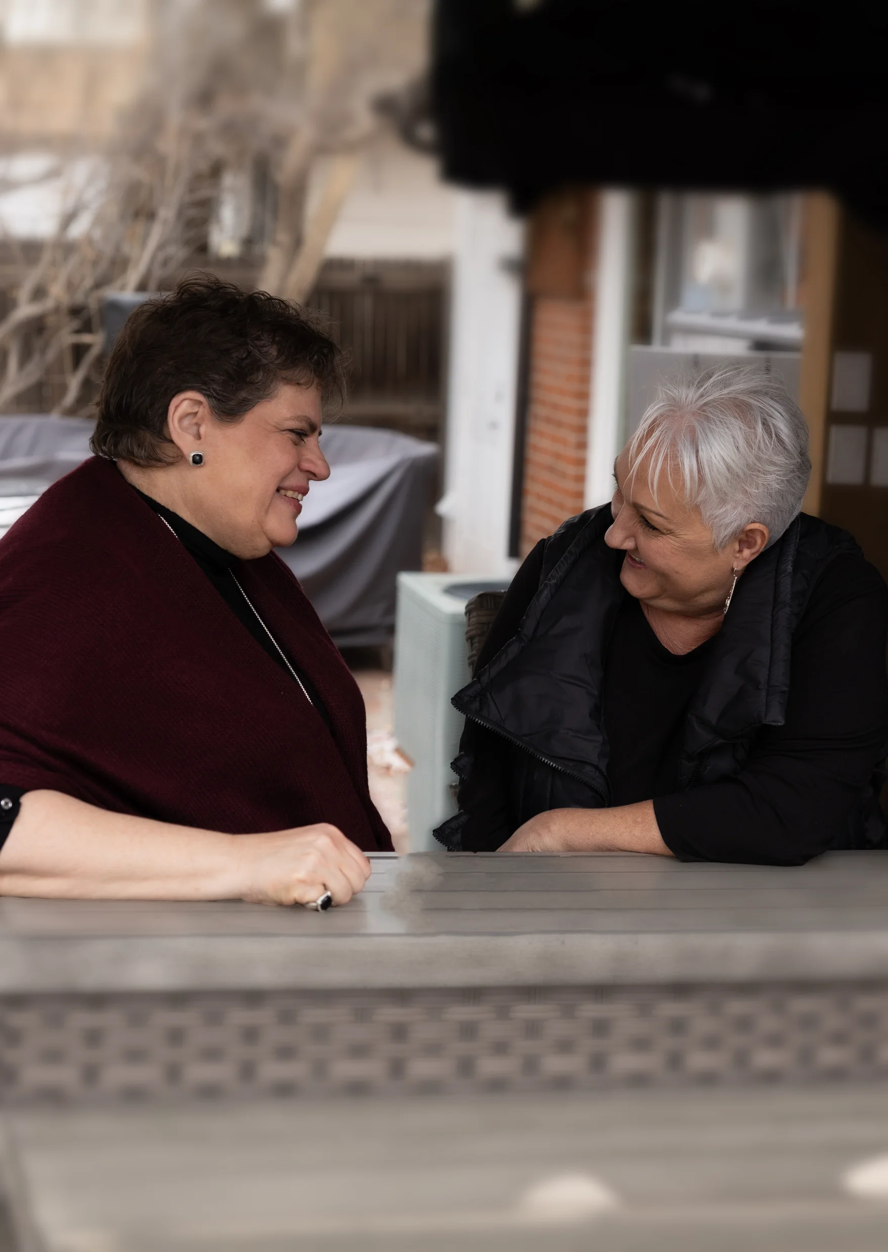 Two older women sitting at an outdoor table facing each other and smiling warmly during conversation.