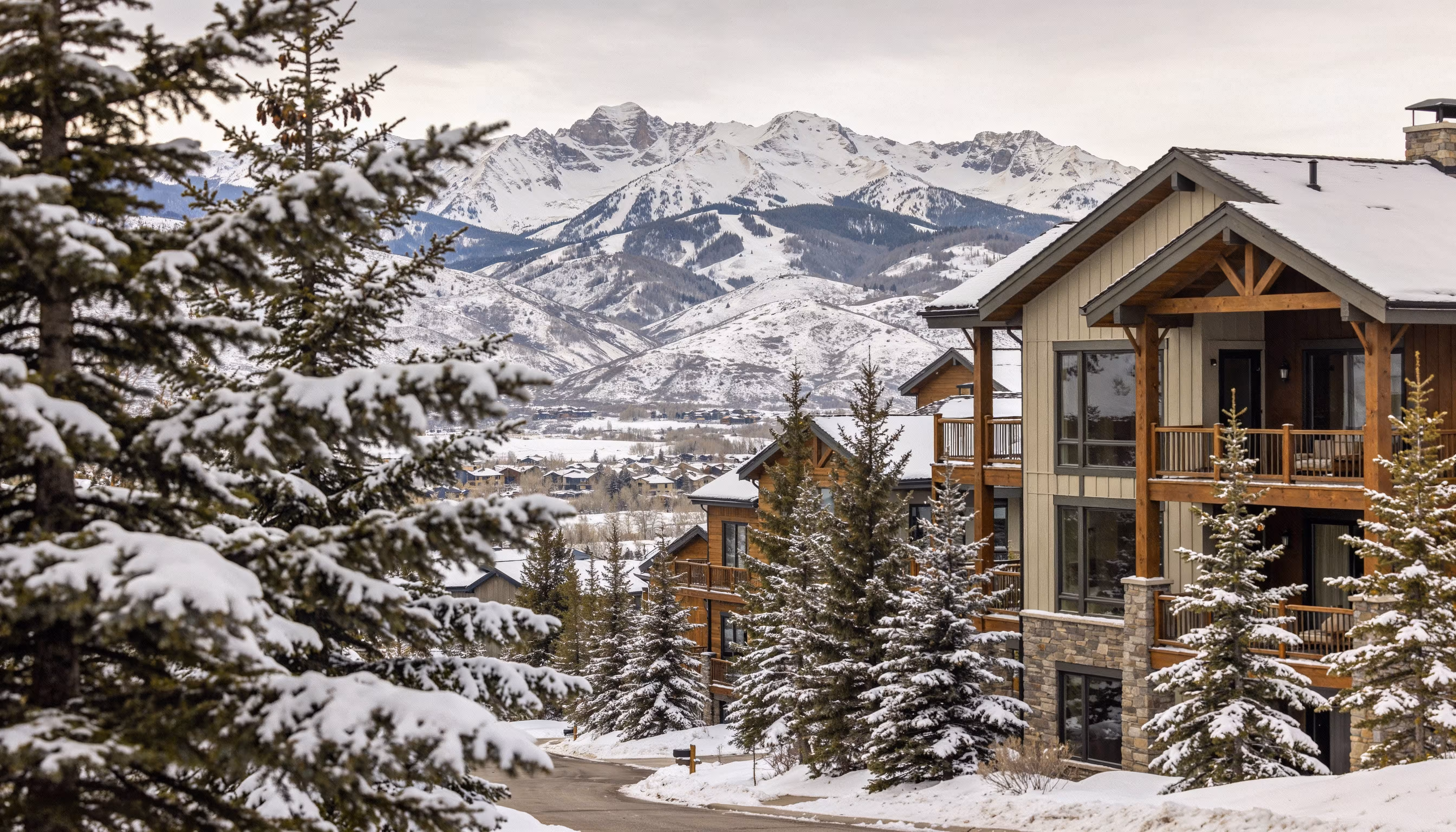 Snow-covered houses and trees on a hillside with snow-capped mountains in the background.