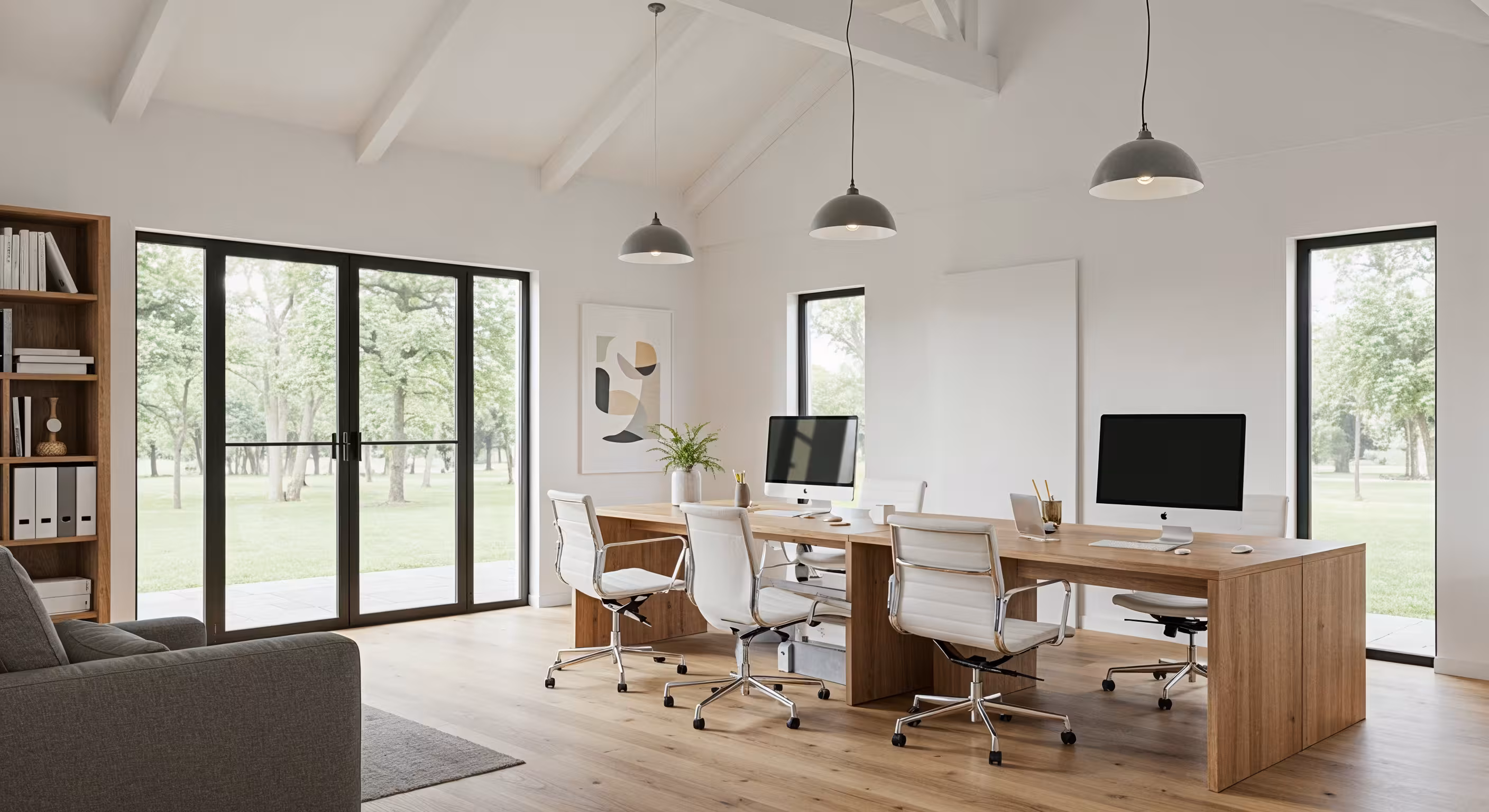 Modern office space with wooden desks, white swivel chairs, desktop computers, and large windows overlooking green trees.