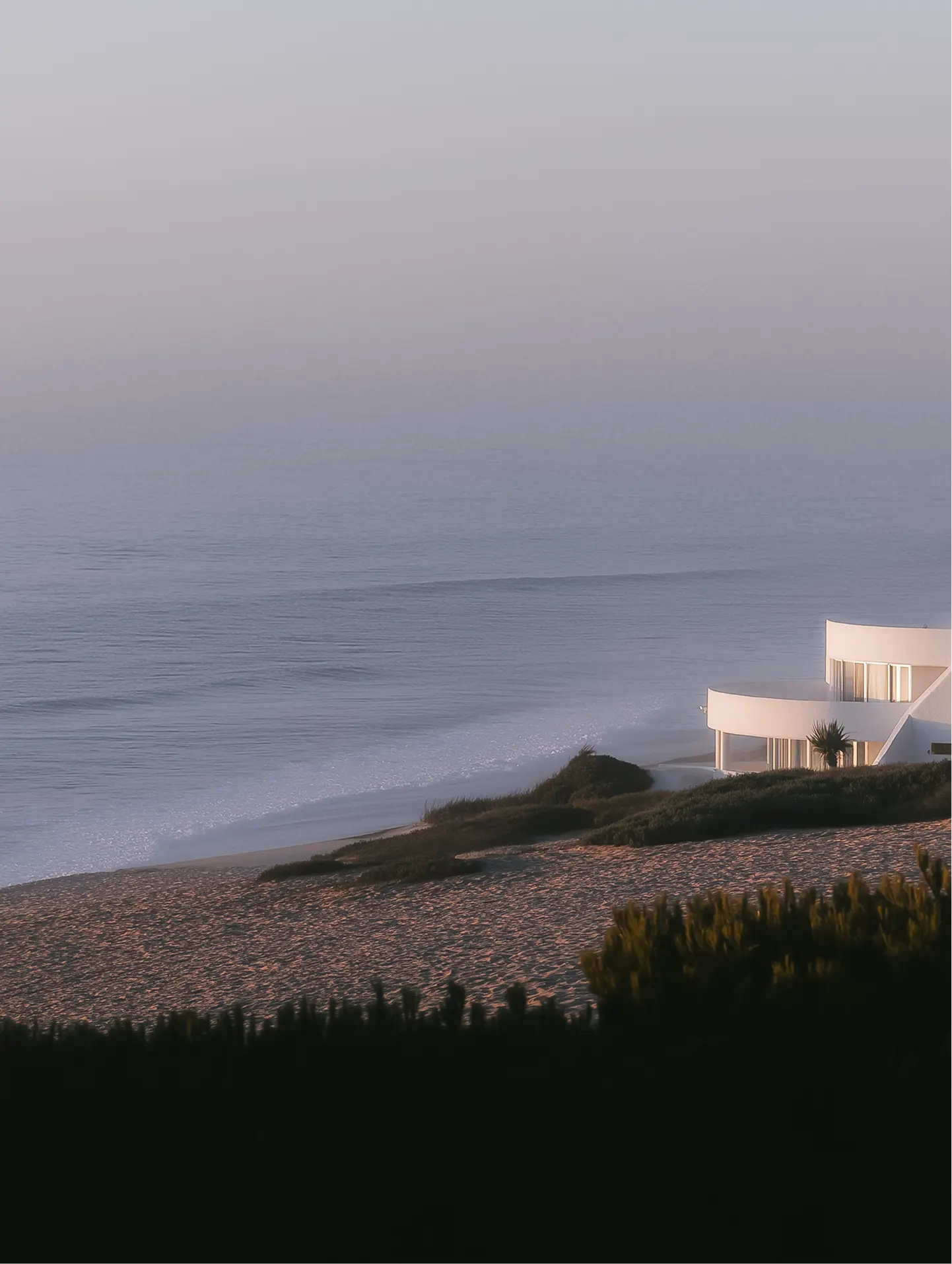 Casa blanca moderna junto a la playa con olas suaves y vegetación en primer plano al atardecer.
