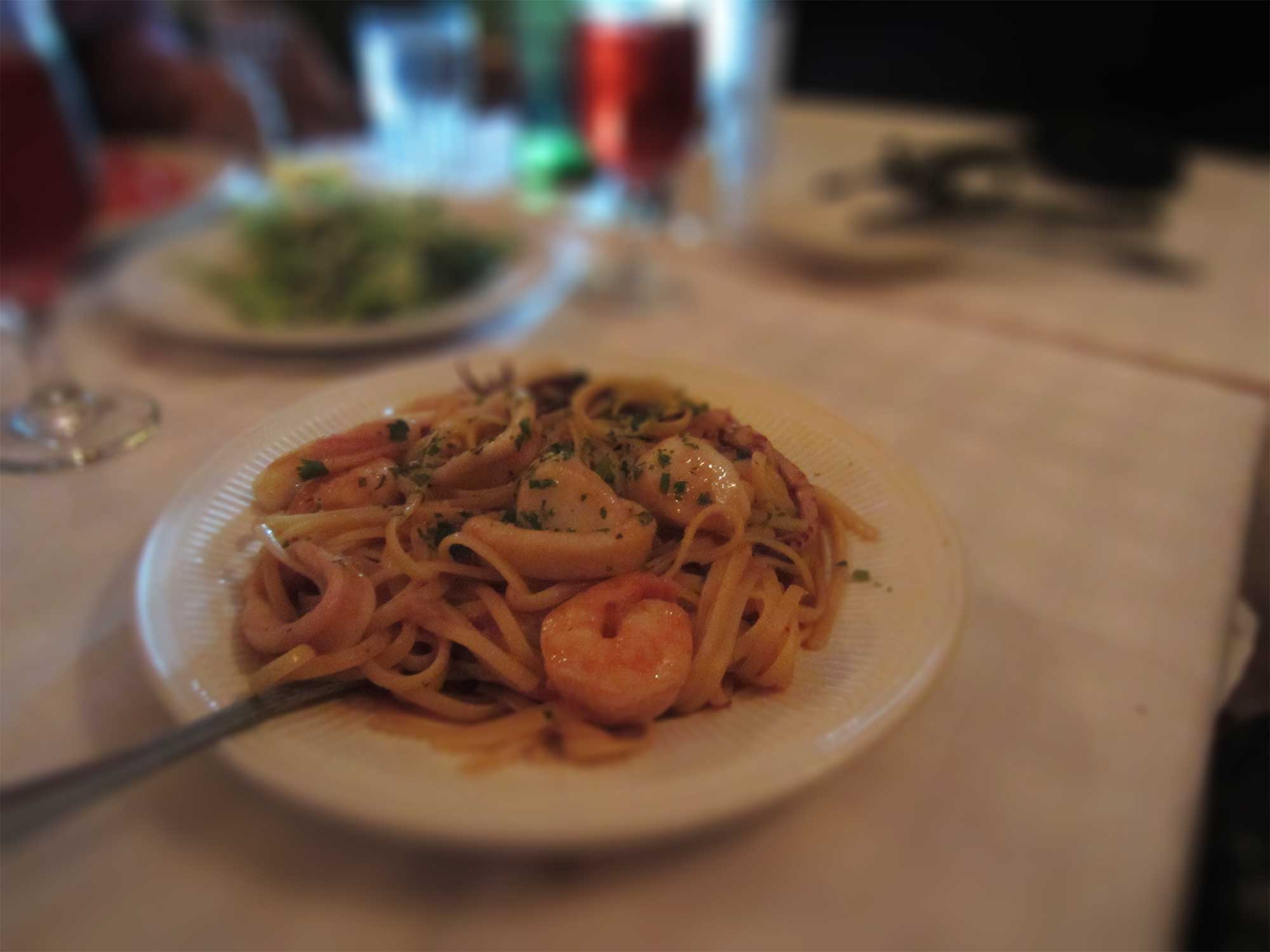 A plate of seafood pasta with shrimp, scallops, and herbs sits on a white tablecloth. The background shows blurred table settings and glasses, creating a cozy dining ambiance.