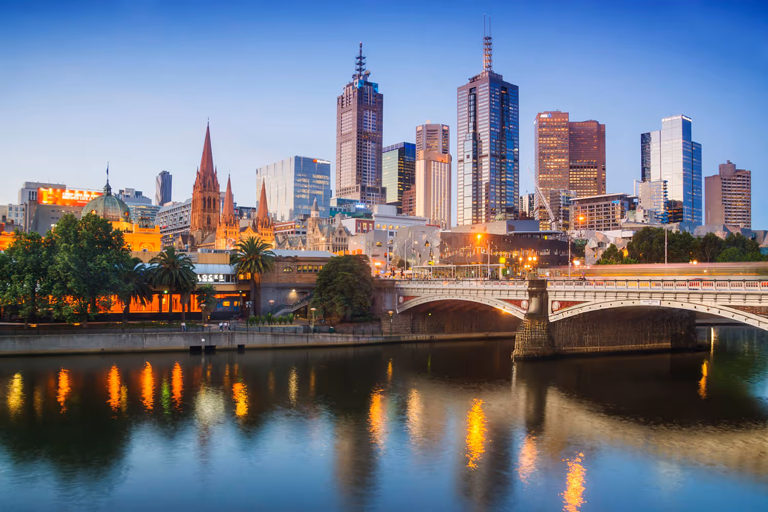 Melbourne city skyline at dusk with illuminated buildings, the Yarra River, and a bridge reflecting lights in the water.