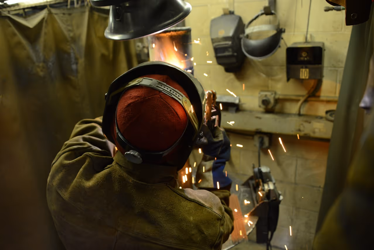 Welder using protective gear while grinding metal, producing sparks inside a workshop