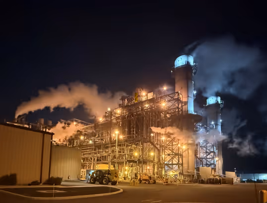 Industrial plant at night with illuminated towers and steam rising against a dark sky.