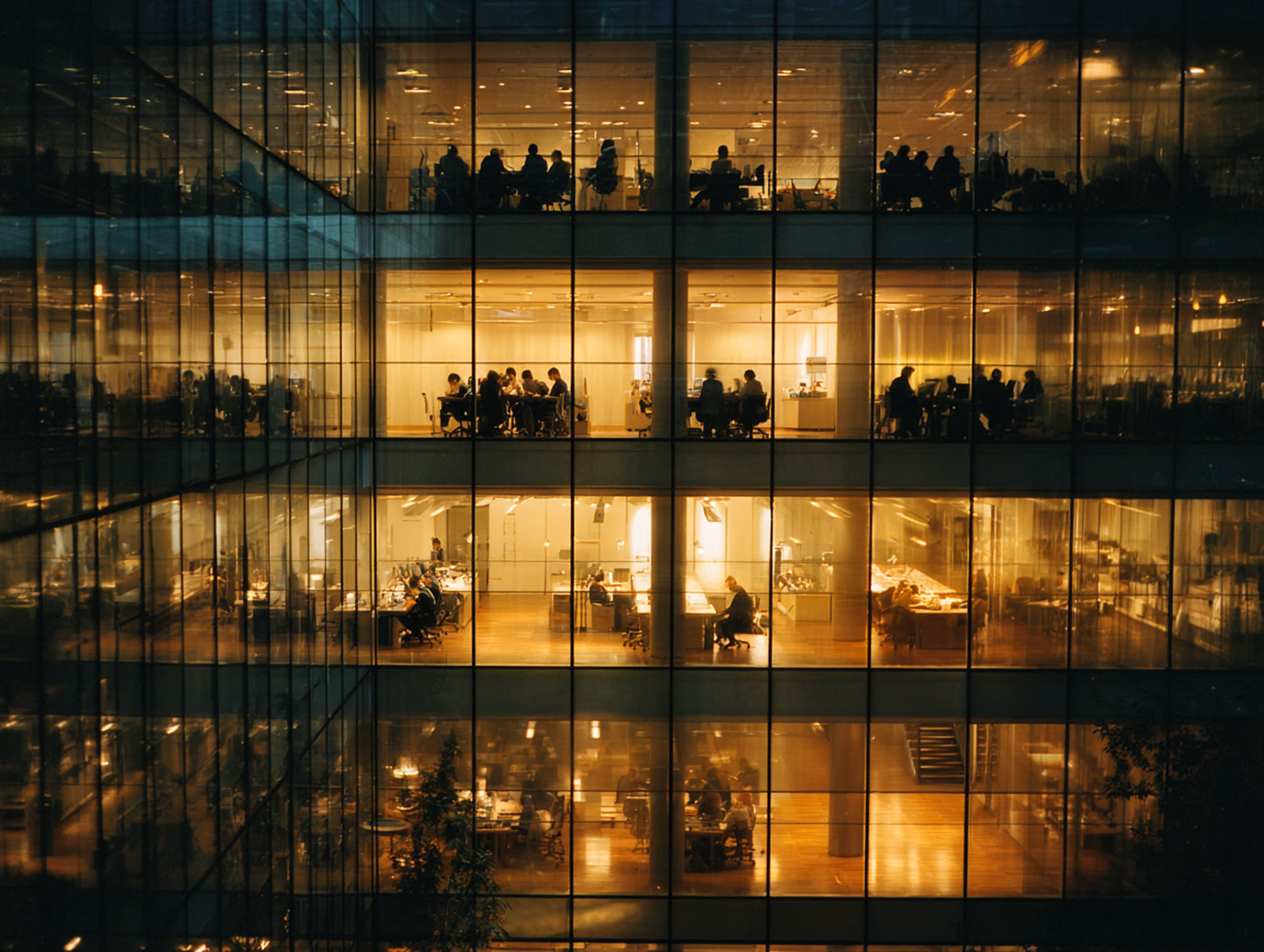 Evening view of a modern office building with multiple floors showing employees working at desks through large glass windows.