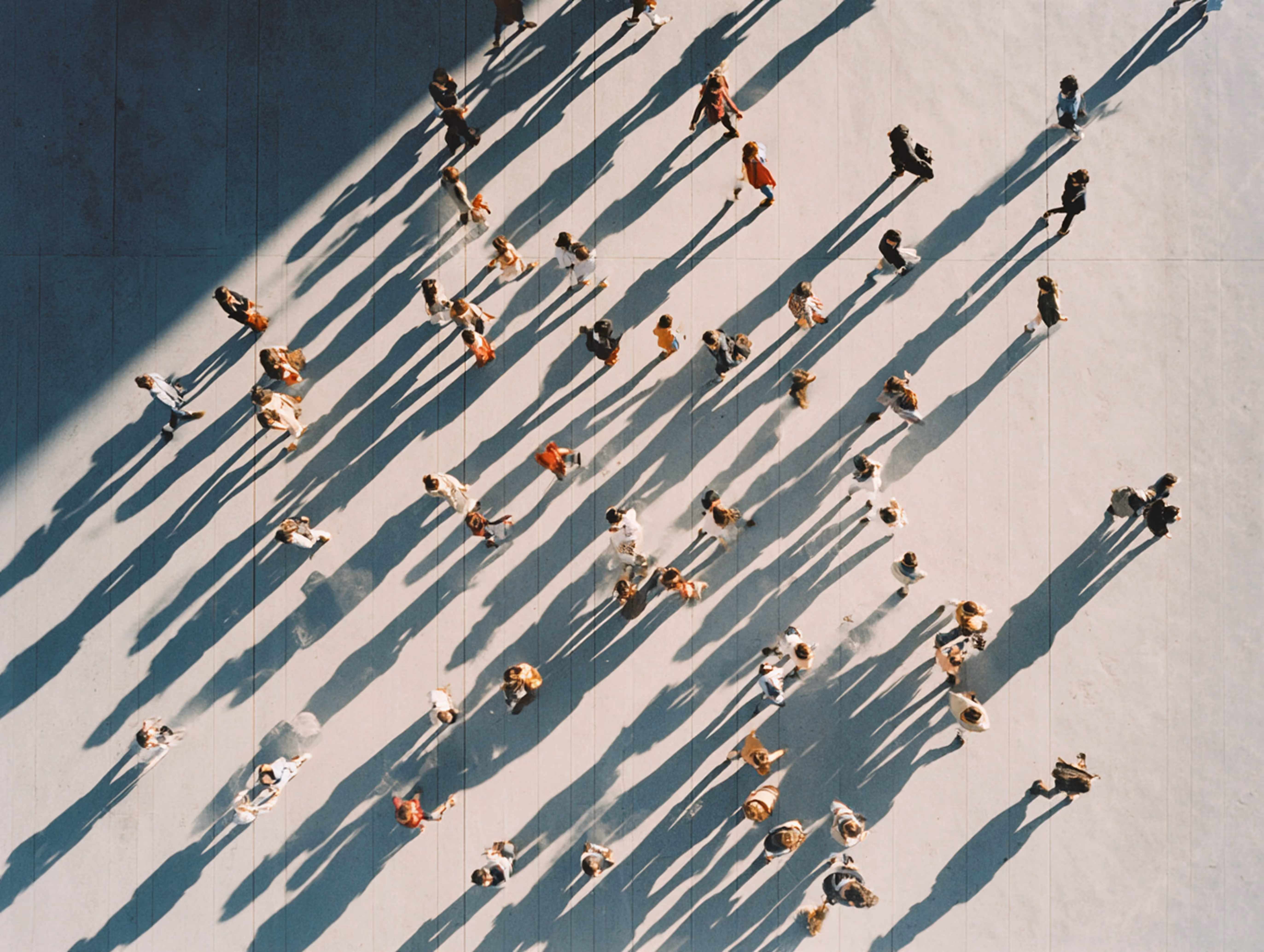 Aerial view of people walking on a sunlit pavement casting long shadows.