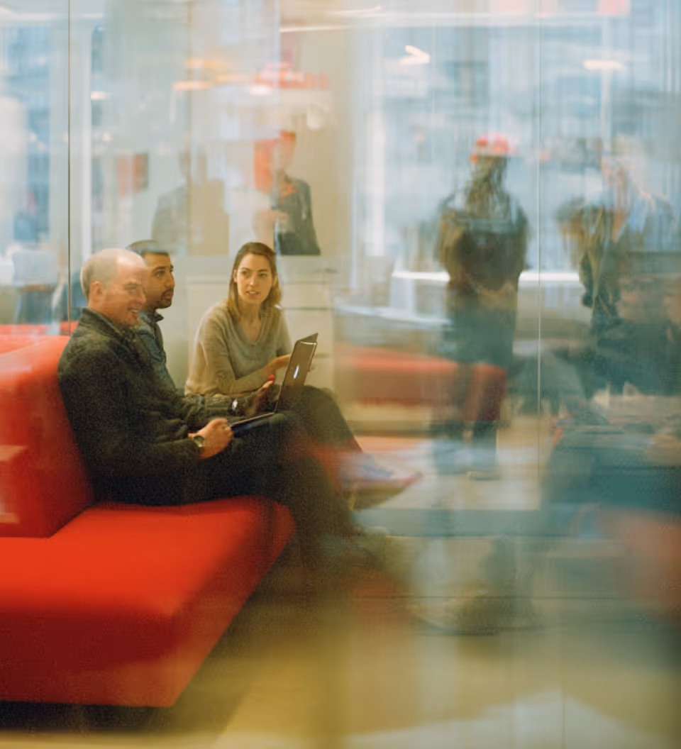Three people sitting on a red couch in an office, two men and one woman using laptop and tablet, with blurred reflections of other people walking by glass walls.