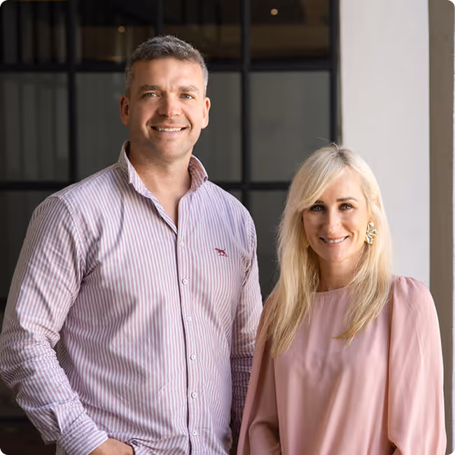 Will smiling in a striped button-up shirt standing next to Nicki with long blonde hair wearing a light pink blouse and gold earrings.