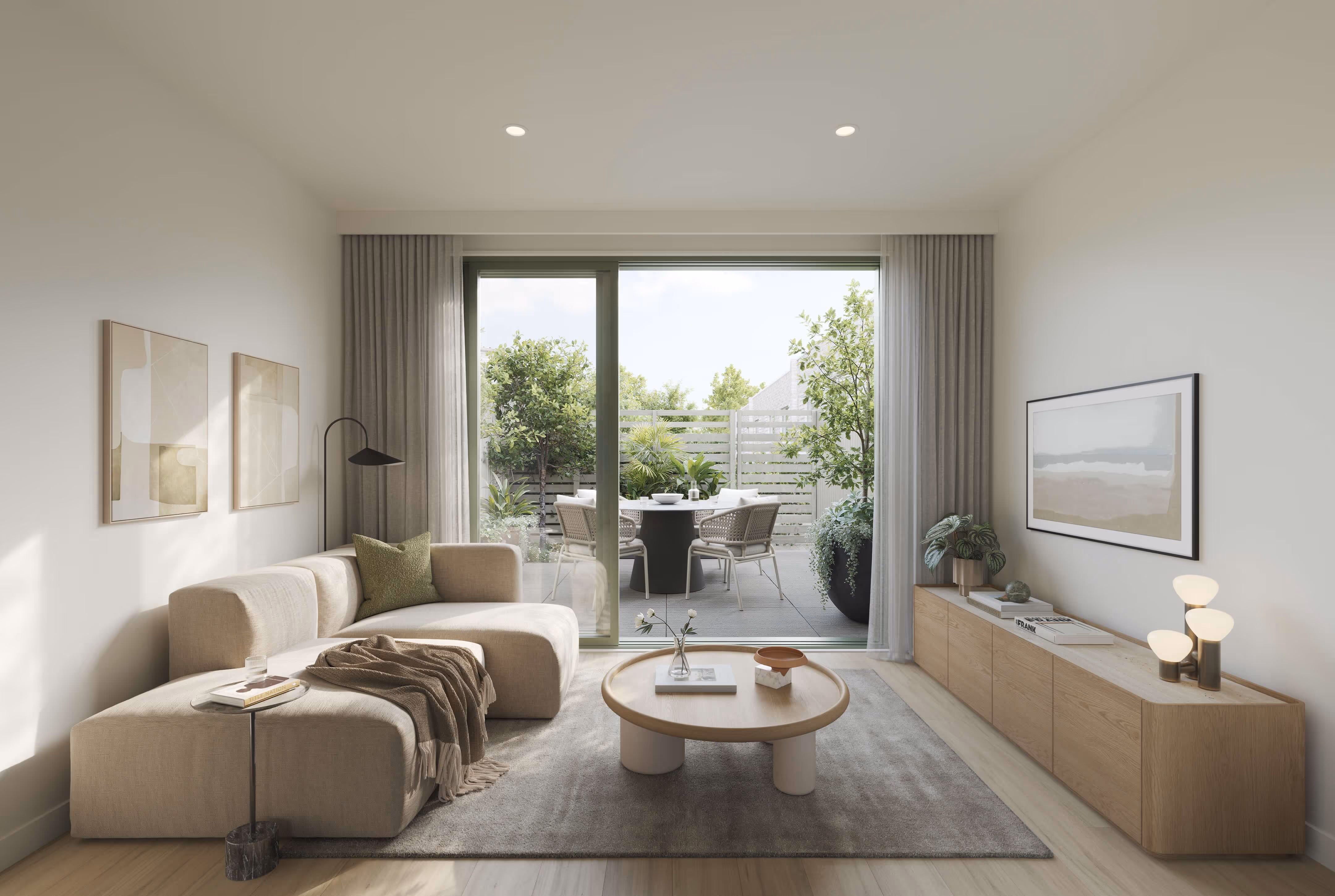 Modern living room with beige sectional sofa, round wooden coffee table, and sliding glass doors opening to a patio with outdoor dining set and greenery.
