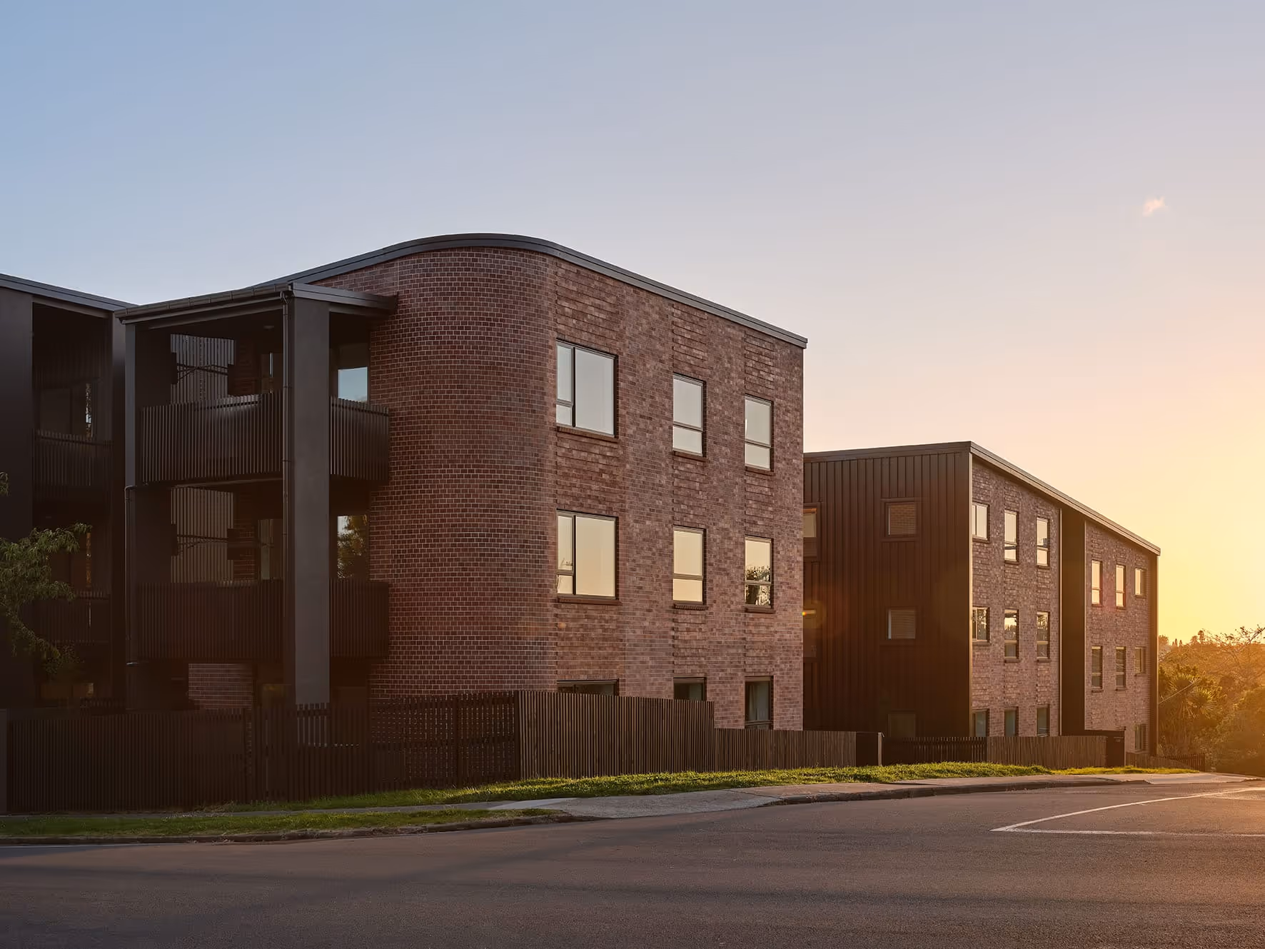 Modern apartment buildings with brick and dark panel exteriors at sunset along a street with a wooden fence.