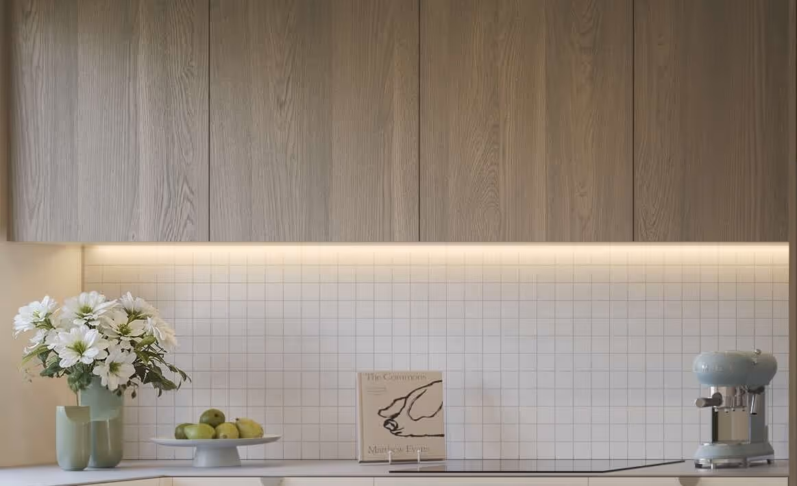 Minimalist kitchen countertop with a bouquet of white flowers in a green vase, a plate of green apples, a cookbook, and a blue espresso machine under wooden cabinets and tiled backsplash with under-cabinet lighting.