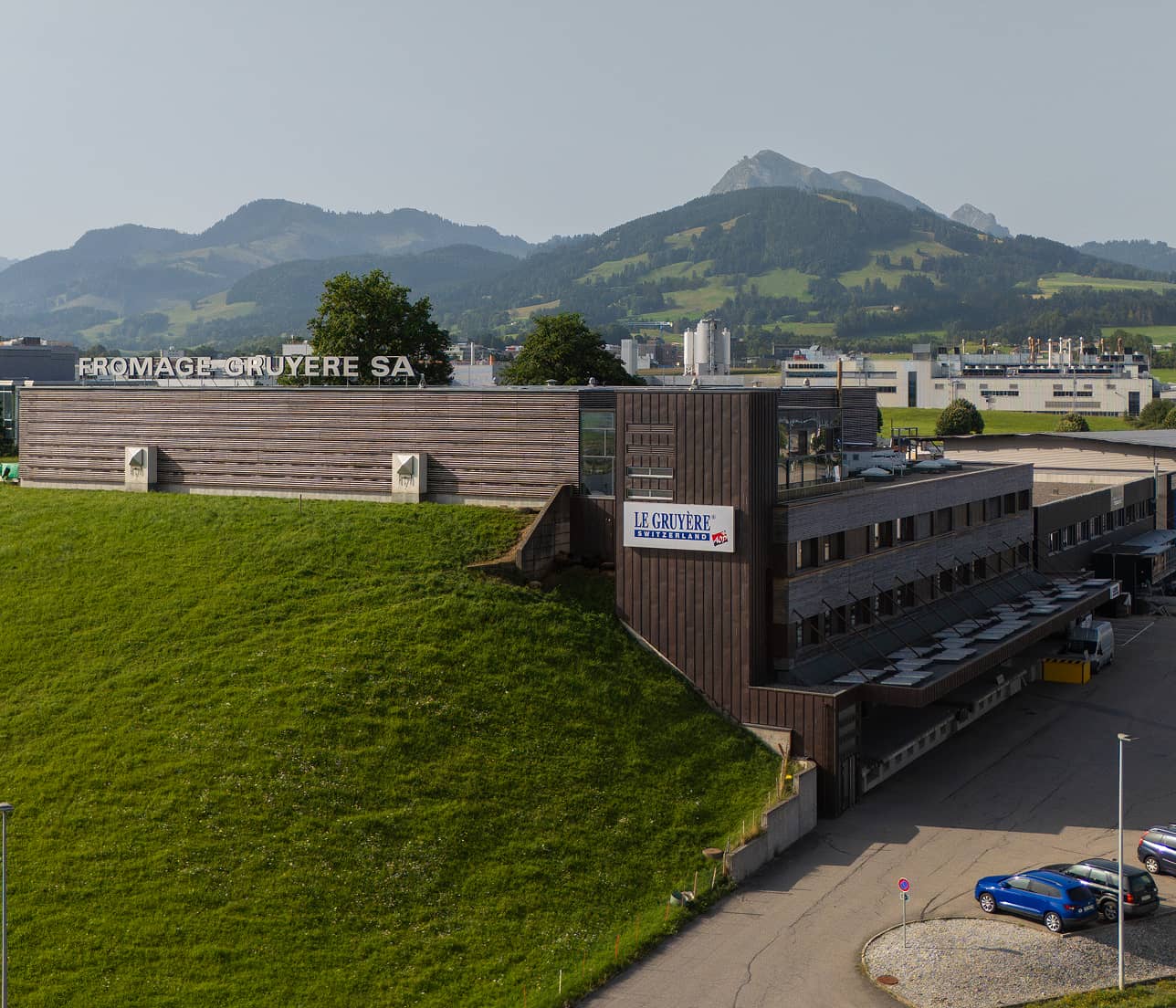Usine Fromage Gruyère SA avec un bâtiment en bois devant une colline verte et des montagnes en arrière-plan.