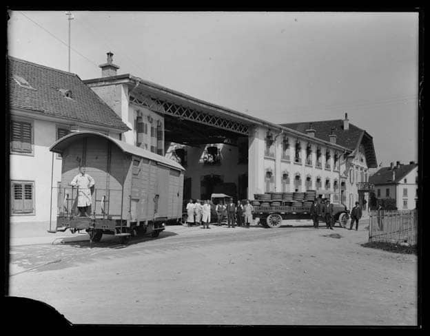Vue en noir et blanc d'un groupe de personnes devant un grand bâtiment industriel avec une charrette chargée de barils et un wagon de train garé.