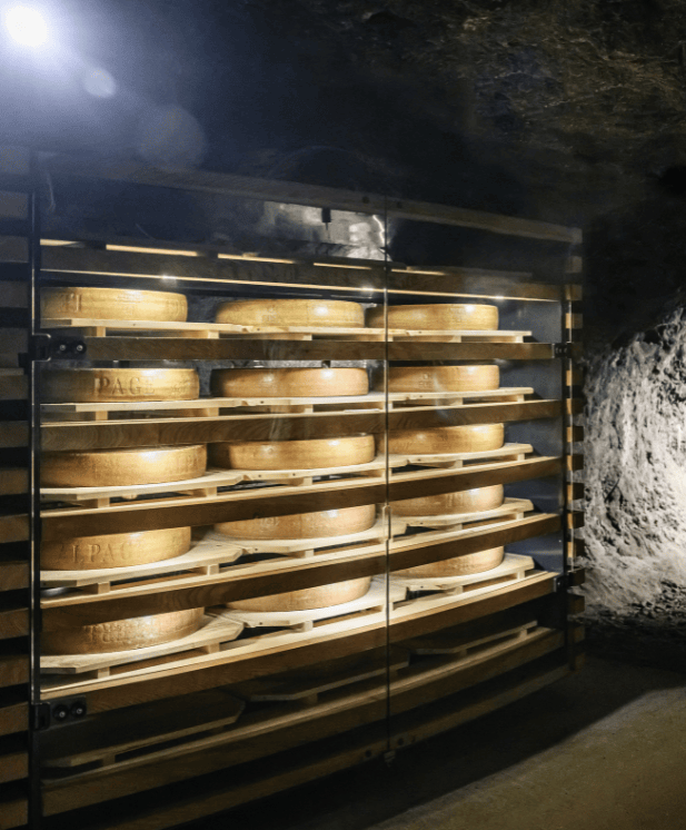 Racks en bois contenant de grandes roues de fromage affinant dans une cave sombre aux murs en pierre.