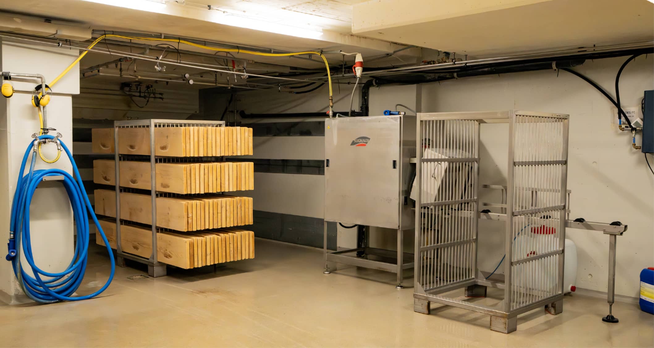 Local industriel avec des blocs de bois empilés sur un chariot en métal, un tuyau bleu enroulé sur un mur à gauche, et des cages métalliques pour équipement.