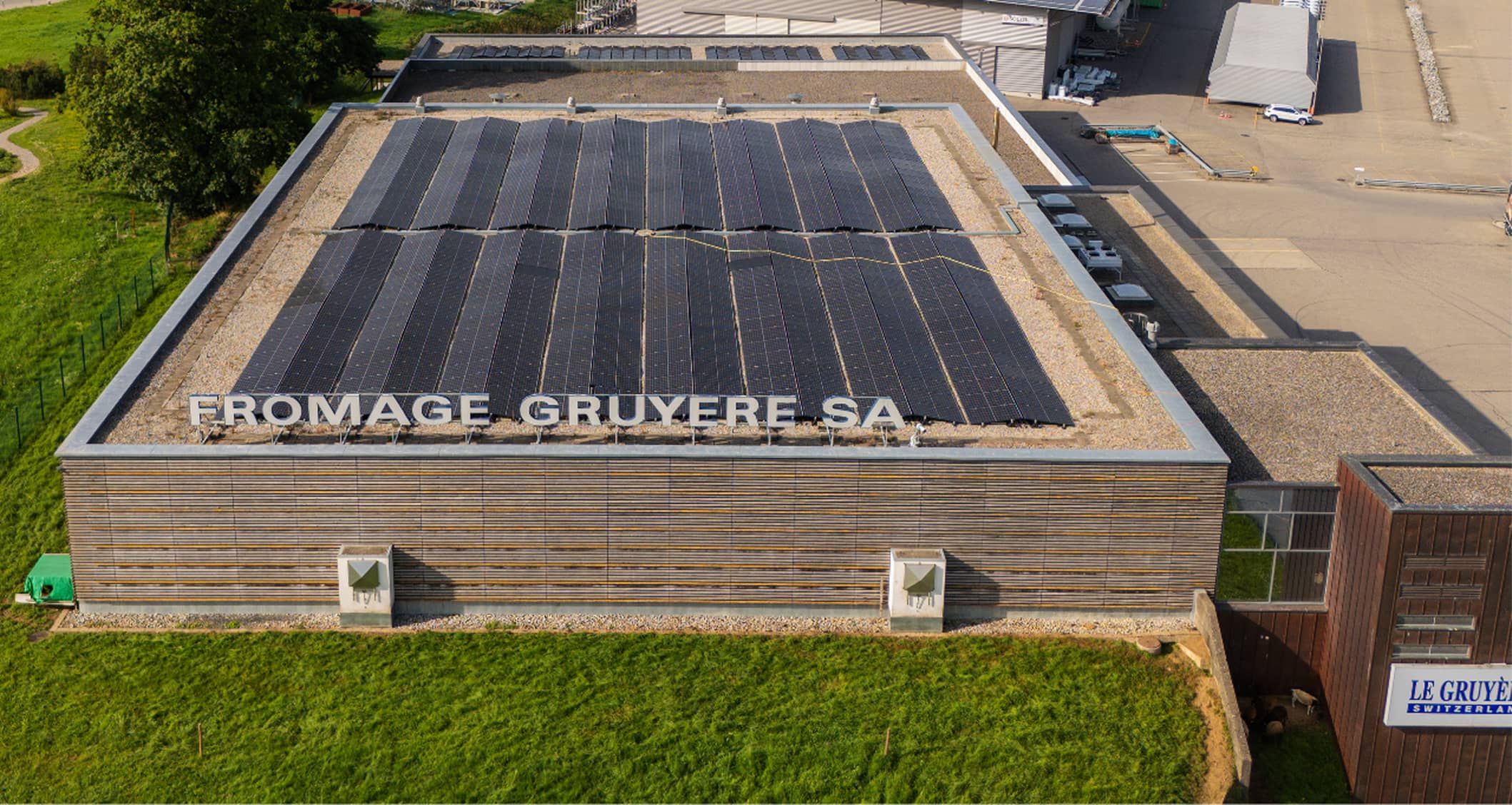 Vue aérienne d'un bâtiment avec des panneaux solaires sur le toit et un grand panneau indiquant FROMAGE GRUYÈRE SA.