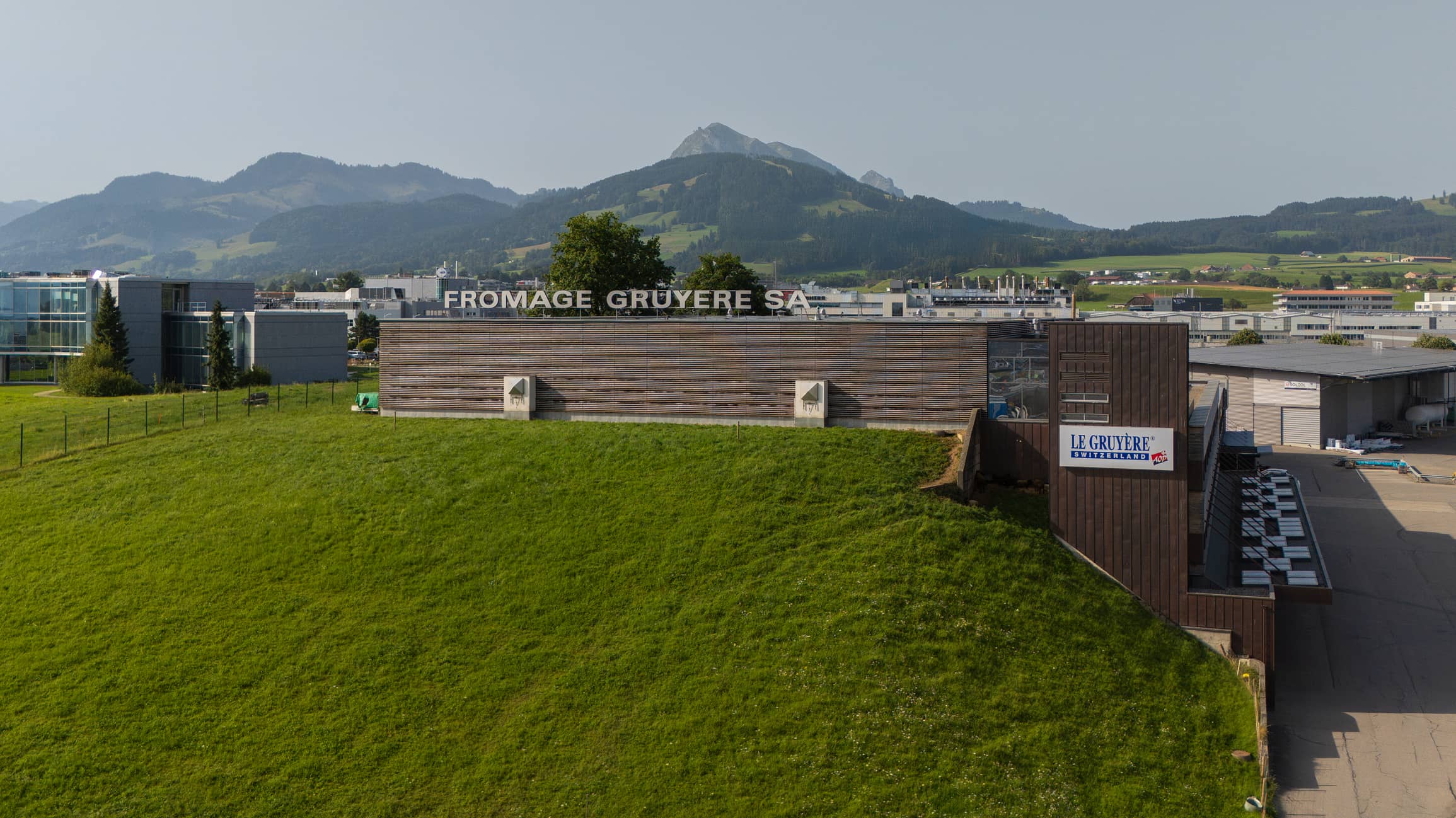 Bâtiment industriel de Fromage Gruyère SA avec des collines vertes et des montagnes en arrière-plan.