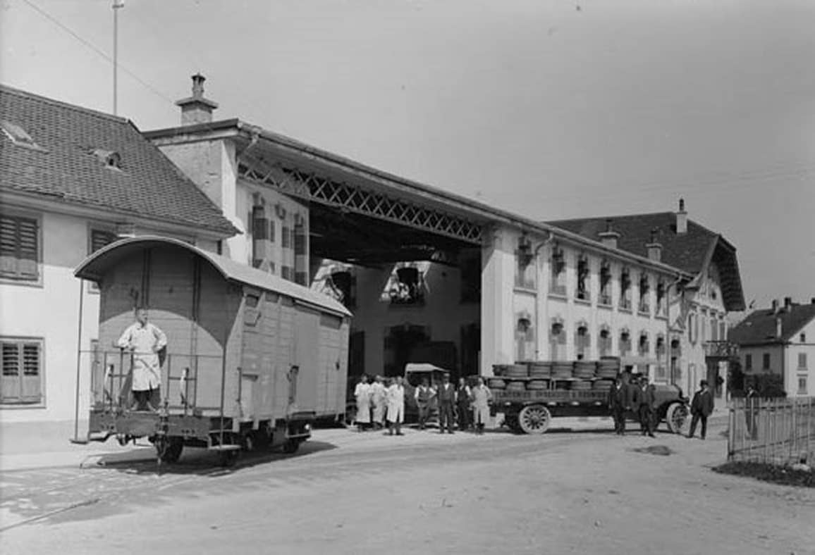 Photo en noir et blanc montrant une scène industrielle avec un wagon de marchandises, plusieurs personnes en tenue de travail, et un camion chargé de barils devant un grand bâtiment.
