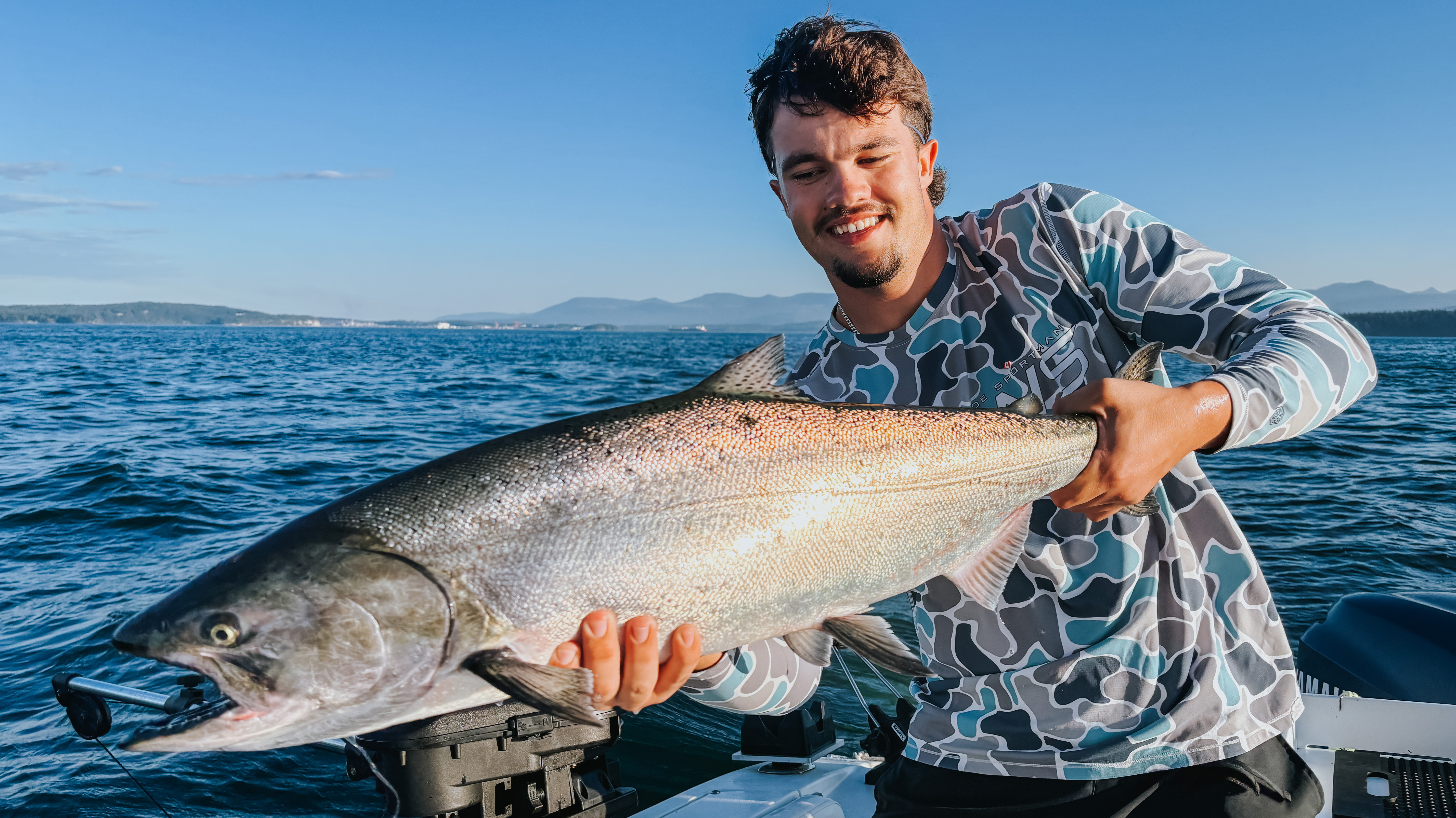 Man smiling and holding a large silver fish on a boat in the ocean with mountains in the background.