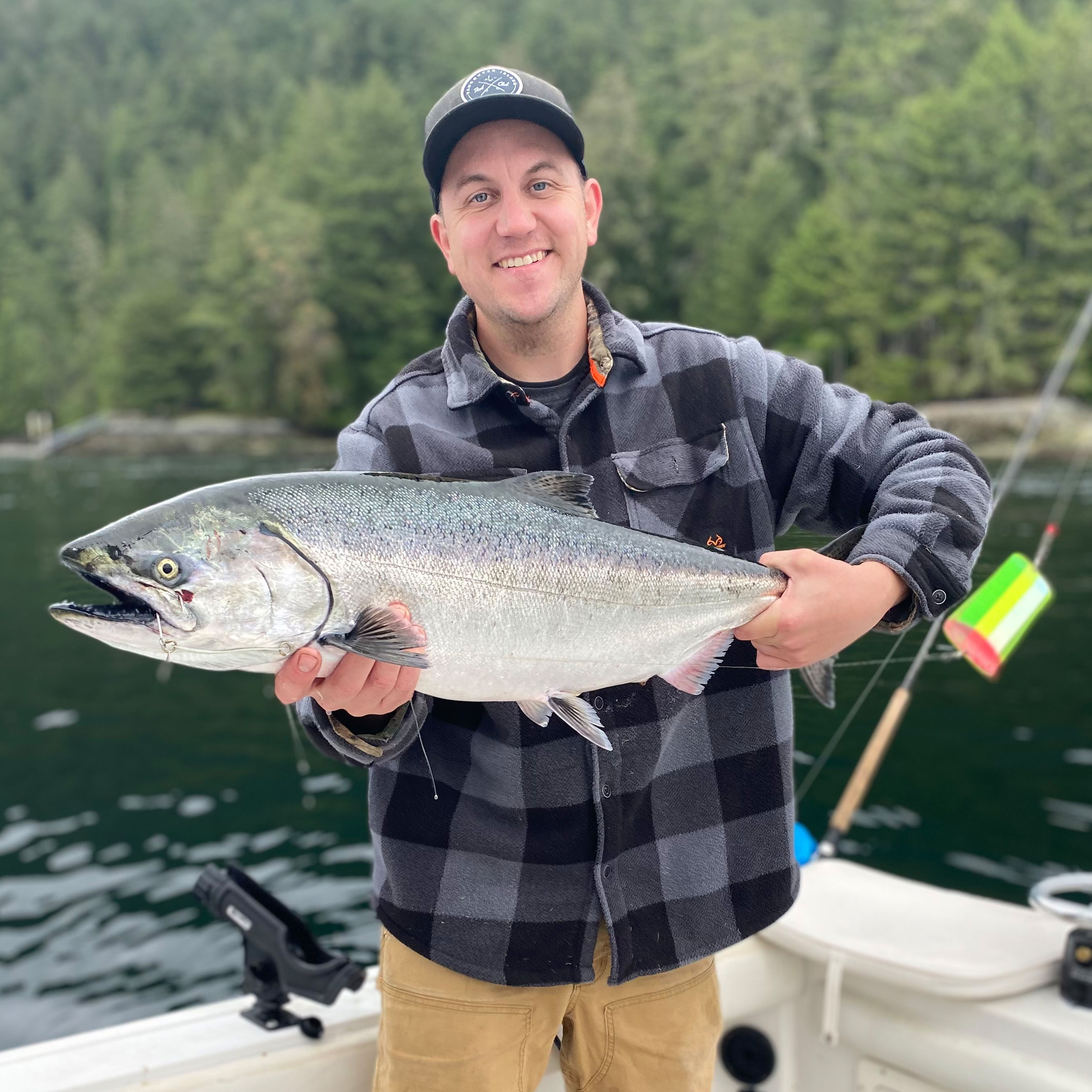 Smiling man in checkered jacket holding a large silver fish on a boat with forest and water in the background.