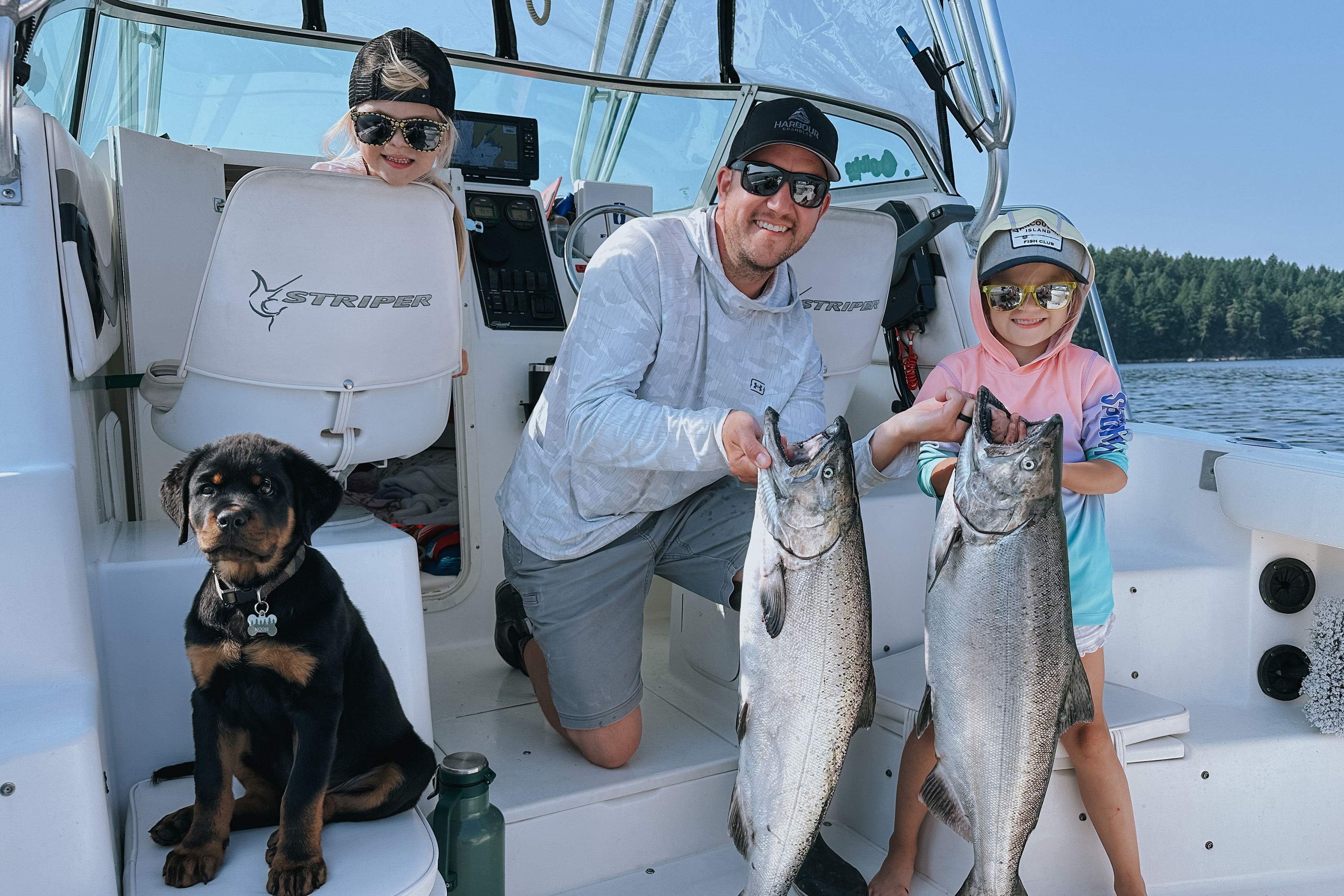 Man and child wearing sunglasses on a boat holding two large fish, with a Rottweiler puppy sitting on a boat seat nearby.