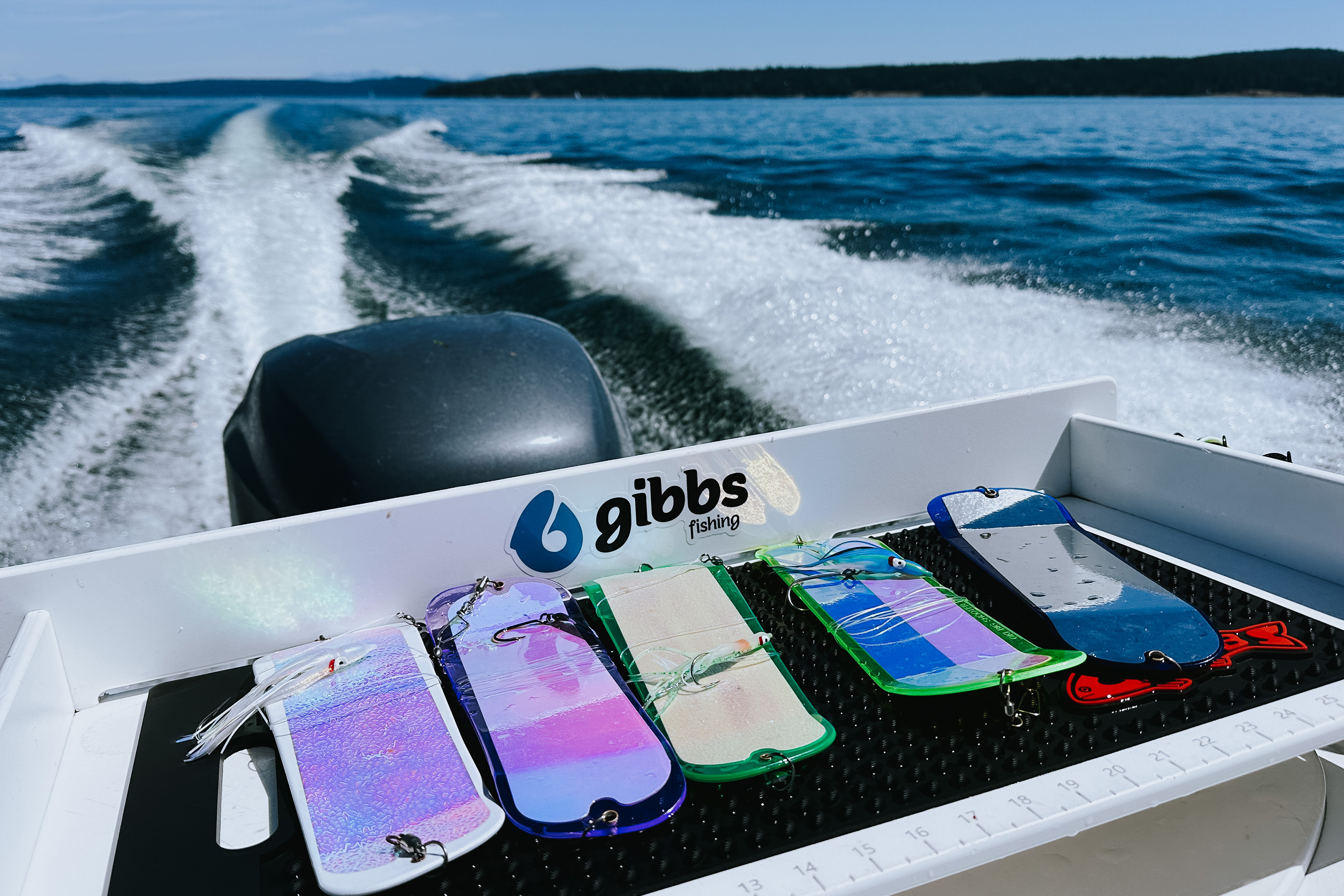 Colorful fishing lures lined up on the back of a boat with water and wake in the background.