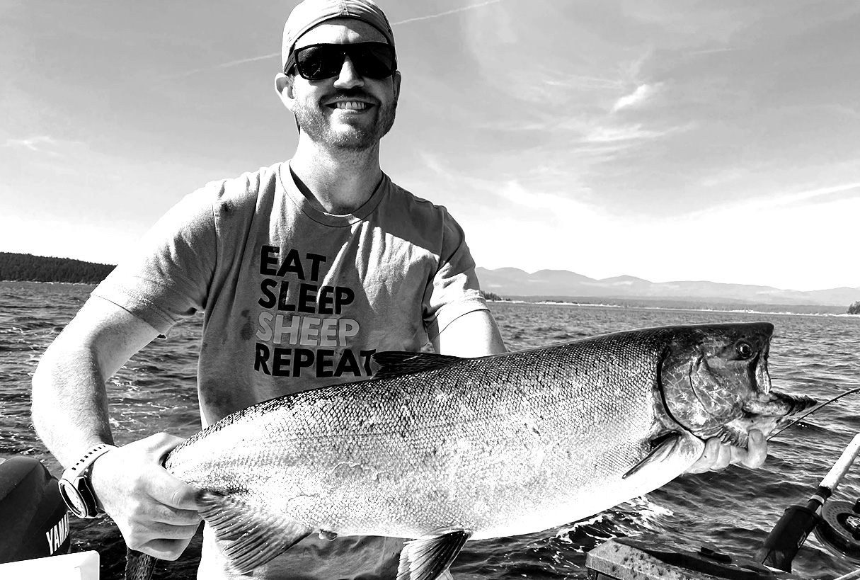 Smiling man wearing sunglasses and a bandana holding a large fish on a boat with mountains in the background.