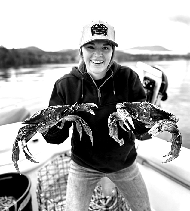 Smiling person wearing a Vancouver Island Fish Club cap holding two large crabs on a boat with water and mountains in the background.