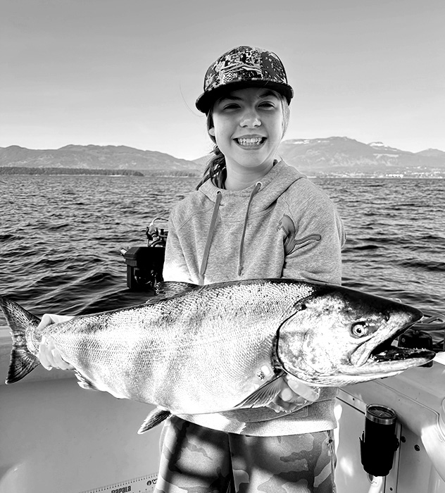 Smiling young person on a boat holding a large fish with mountains and water in the background.
